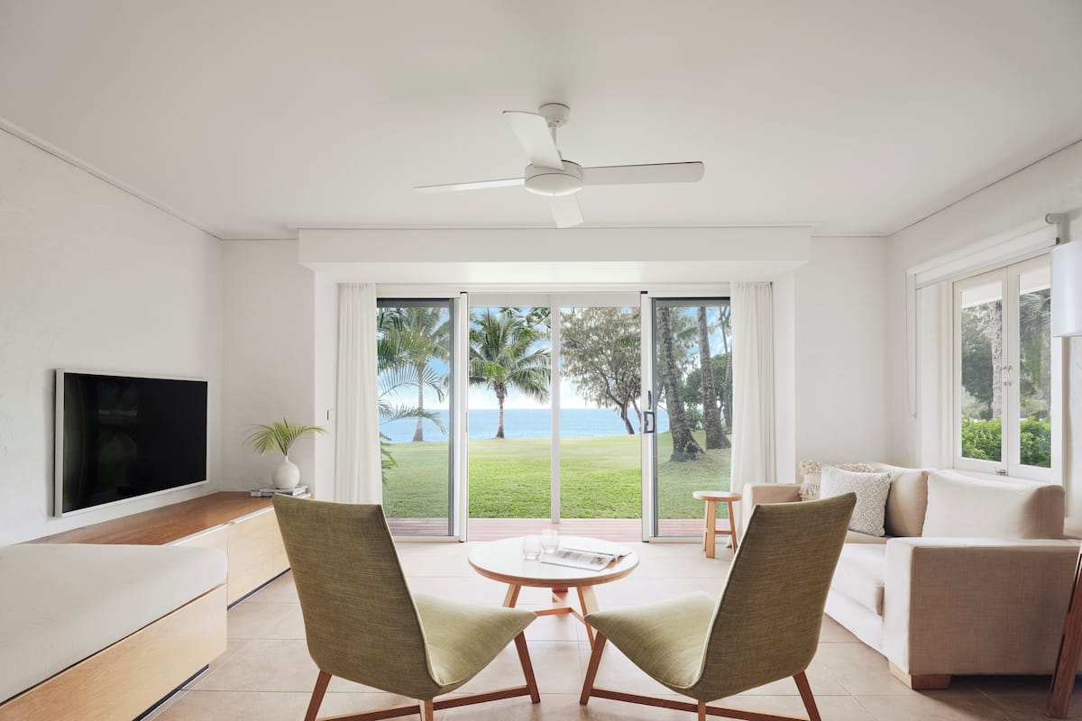 Una sala de estar moderna con un televisor de pantalla plana, dos sillones, un sofá y un ventilador de techo. Amplias puertas correderas de vidrio dan a un área verde con palmeras y vistas al océano.