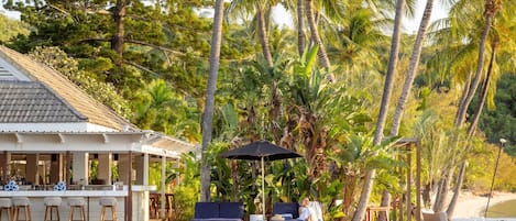 A waterfront bar with outdoor seating, surrounded by palm trees and a pool.