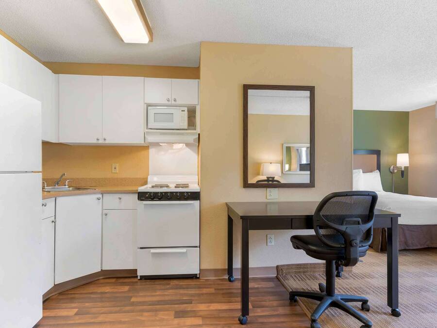 A compact hotel room with a kitchenette, a bed, a desk with a chair, and a mirror.