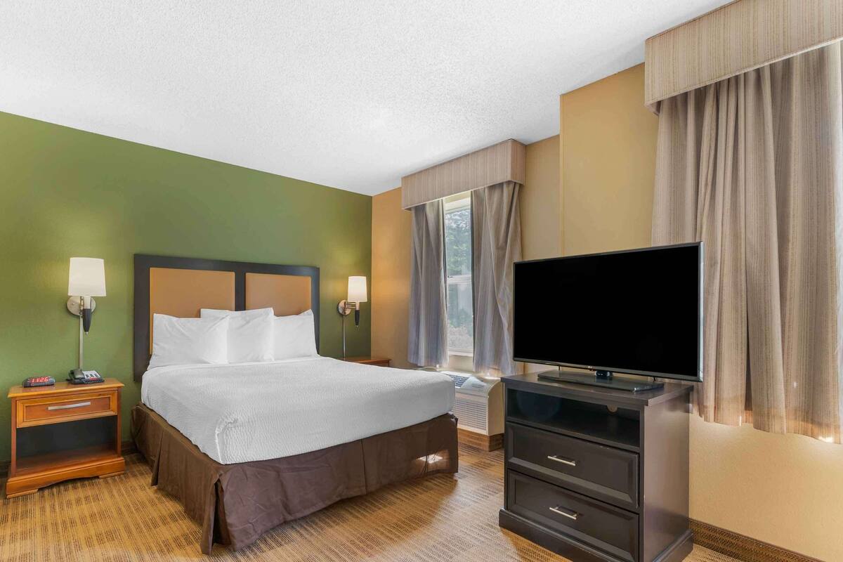 Una habitación de hotel con cama, lámparas de noche, televisor, cómoda y ventana con cortinas.