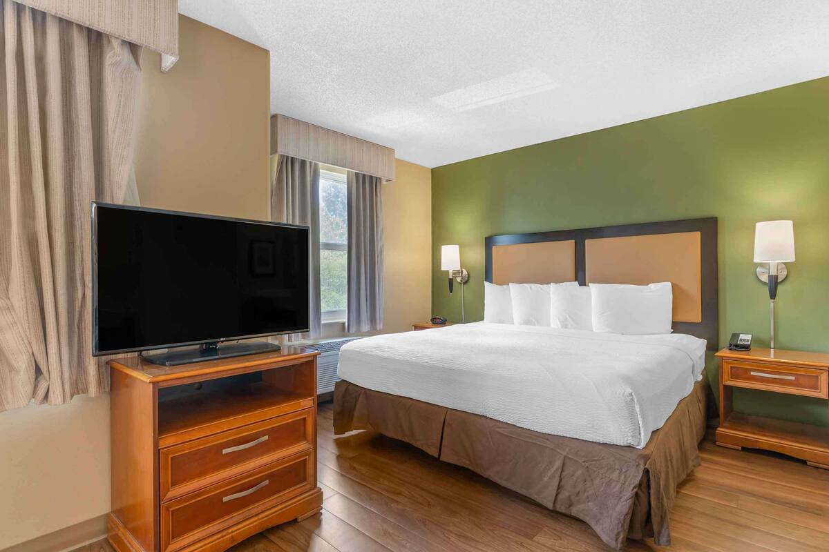 Habitación de hotel con una cama grande, un televisor de pantalla plana sobre un tocador de madera, dos lámparas de noche y una ventana con cortinas.
