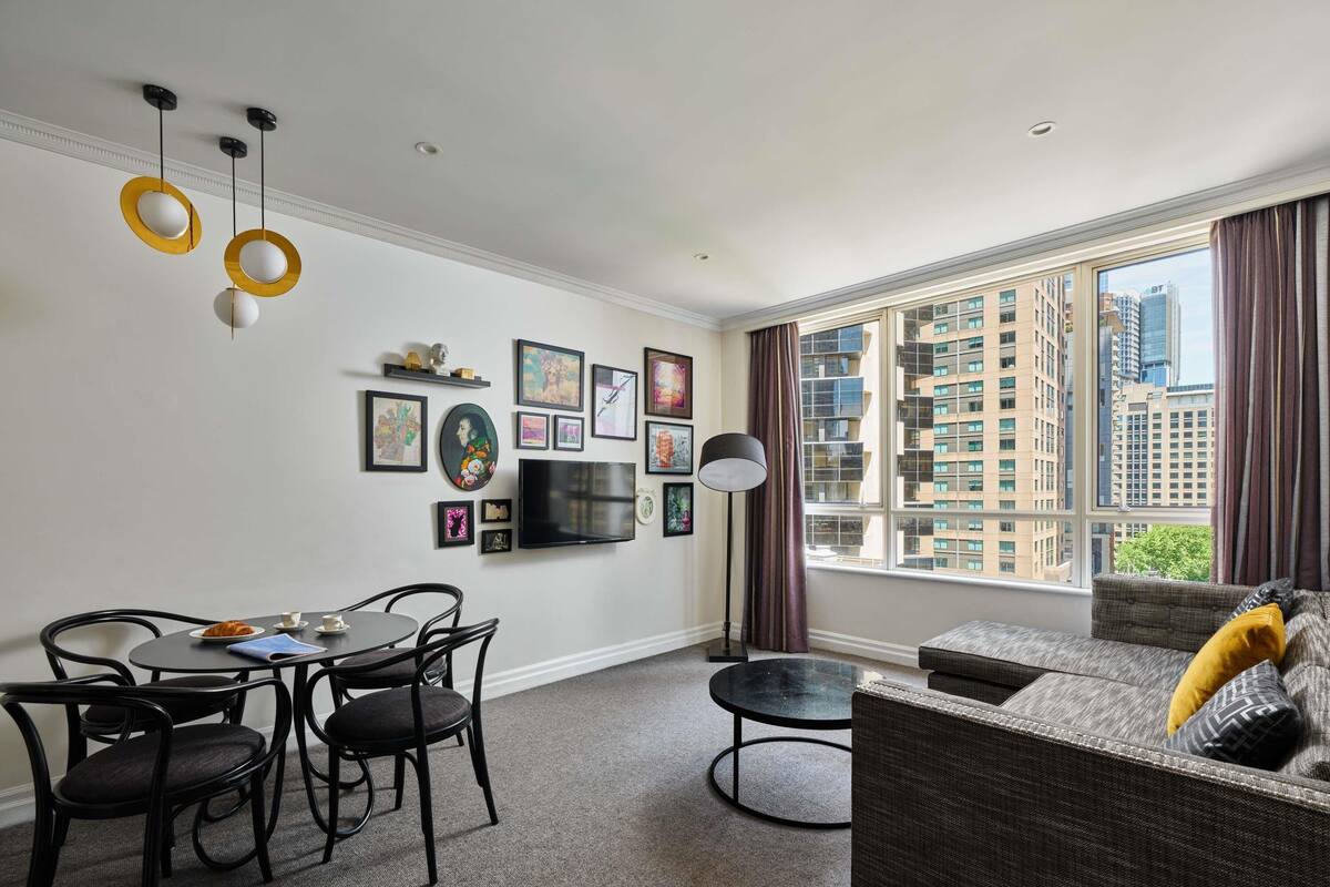 A modern living room with a dining area, large window with city view, and artwork on the wall.
