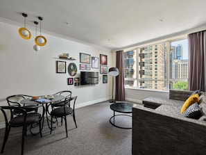 A modern living room with a dining area, large window with city view, and artwork on the wall.