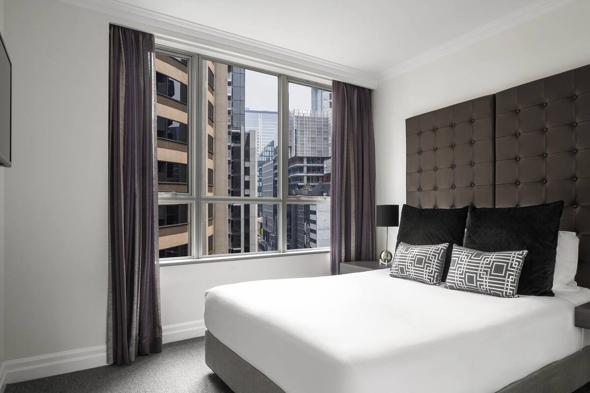 A hotel room with a large bed, a headboard, a window with curtains, and a view of buildings.