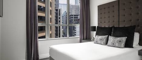 A hotel room with a large bed, a headboard, a window with curtains, and a view of buildings.