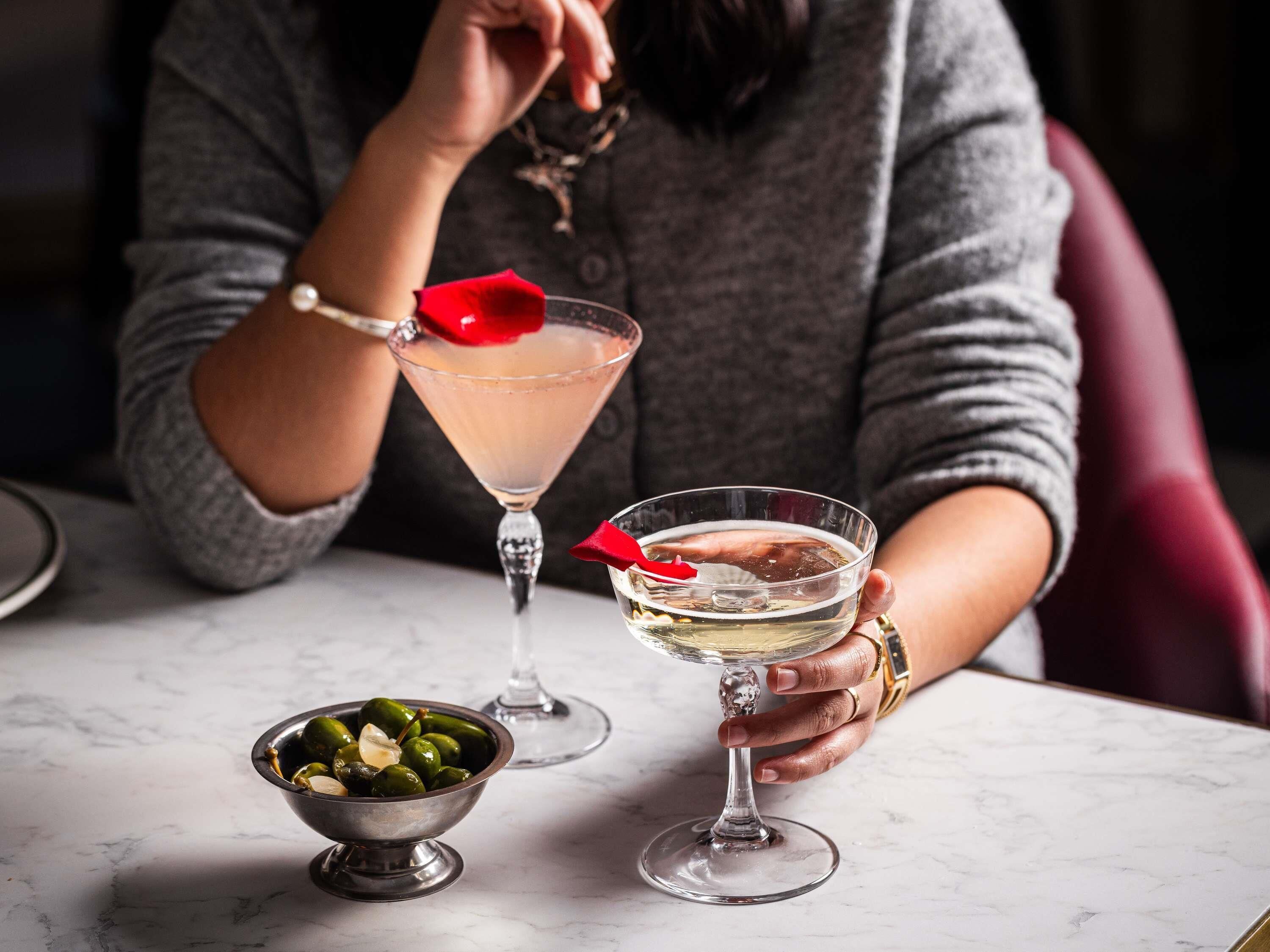 A person holding two glasses of a clear beverage, a bowl of olives, and a martini glass with a red rose on the table.