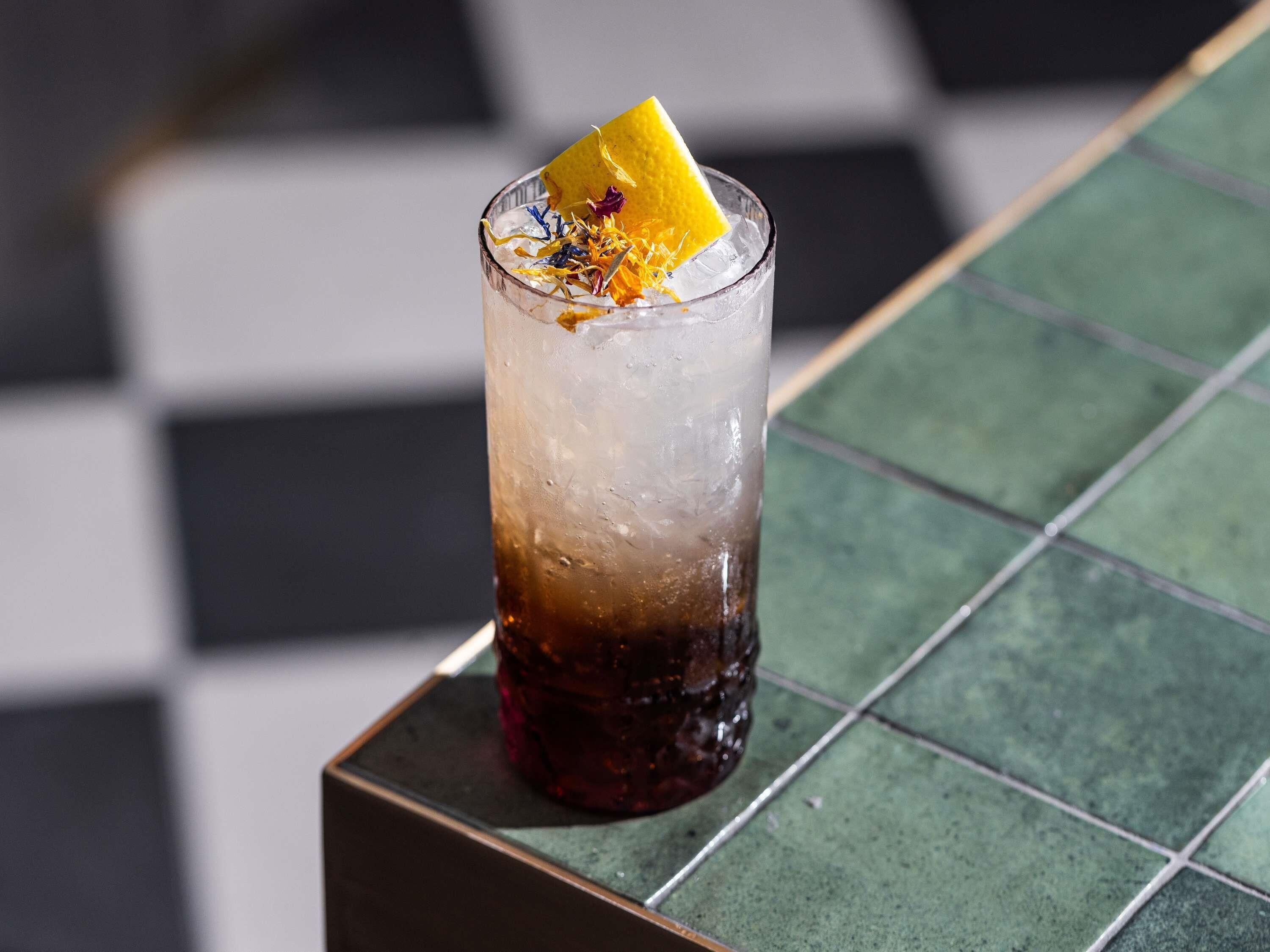 A glass with a dark liquid, ice, and a yellow slice on a tiled surface.