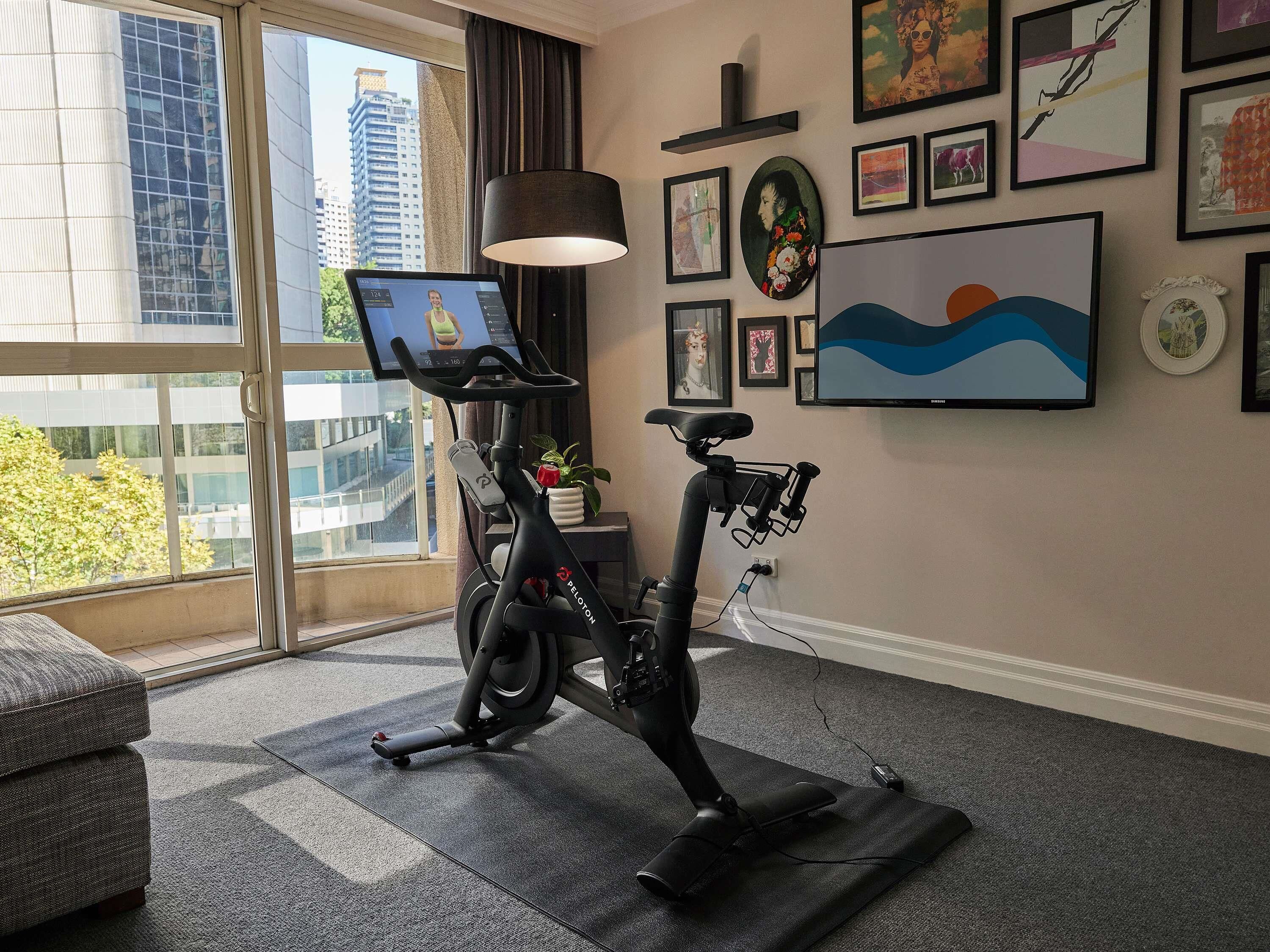 A room with a stationary bike, a laptop on a stand, a large window with a view of buildings, and various framed artworks on the wall.