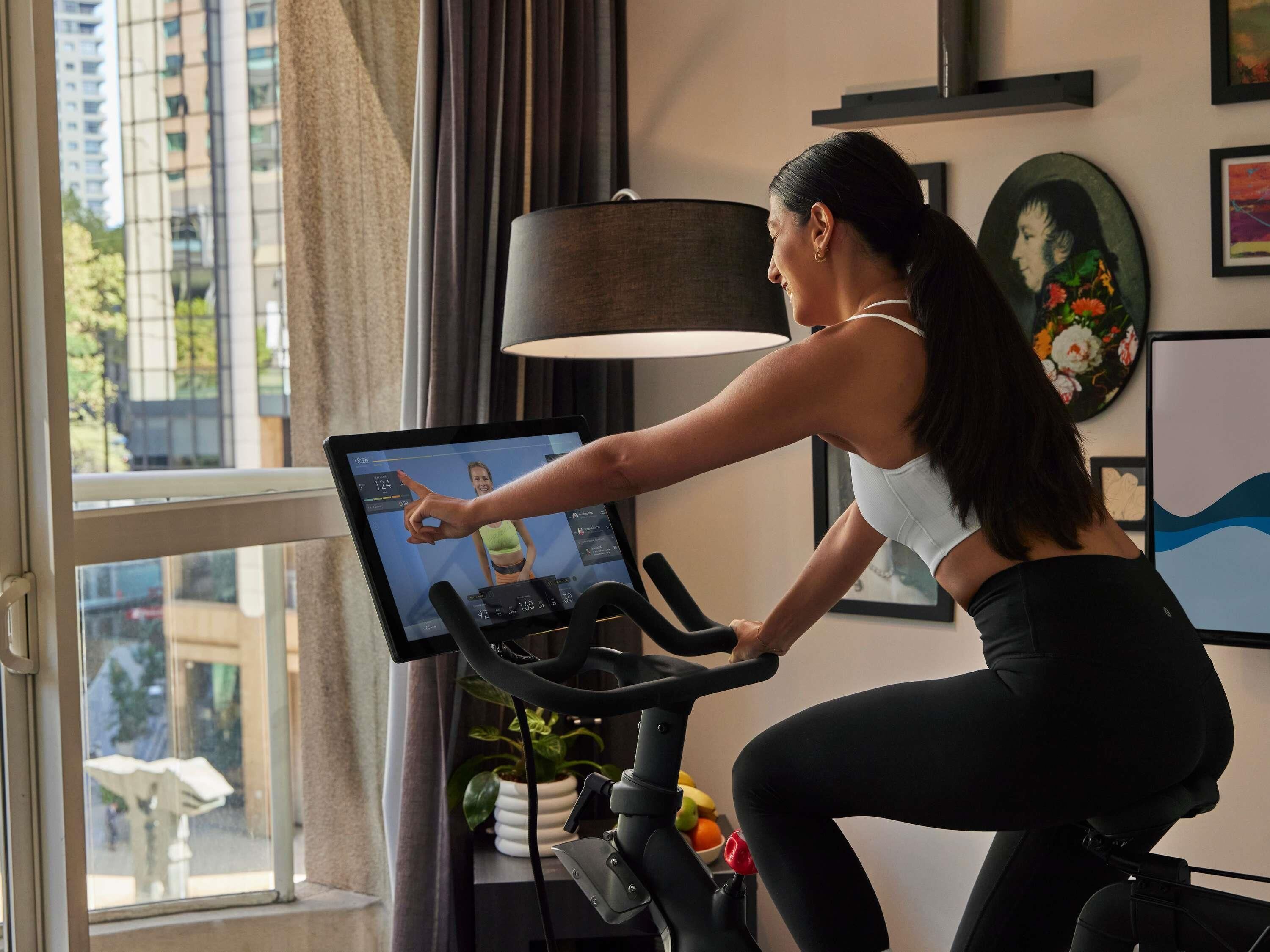 A person on an exercise bike with a digital display, a window with curtains, and a wall with framed artwork.