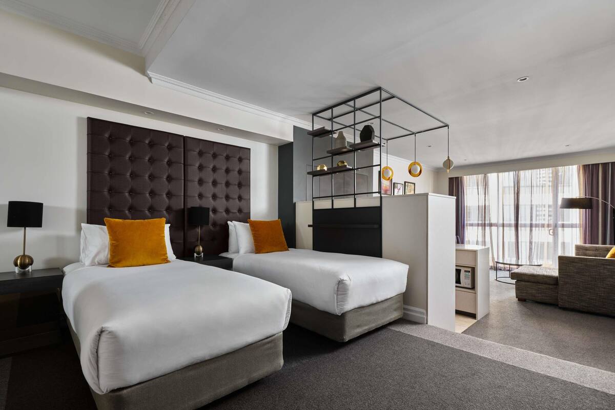 A hotel room with two beds, a large tufted headboard, a desk, and a view through large windows.