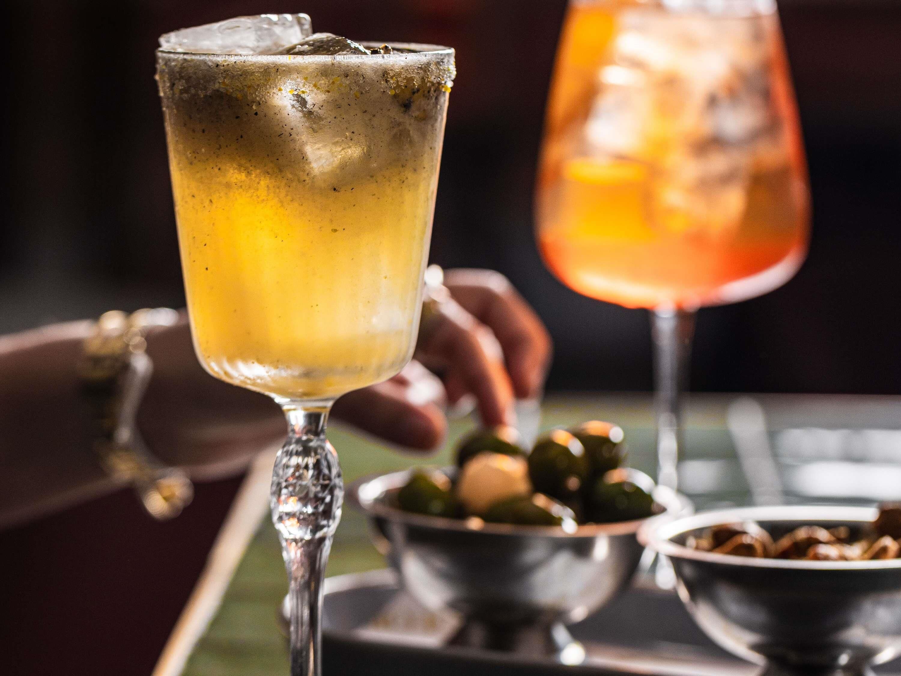 Un bar con un bicchiere di una bevanda gialla, uno sfondo sfocato e una ciotola di olive.
