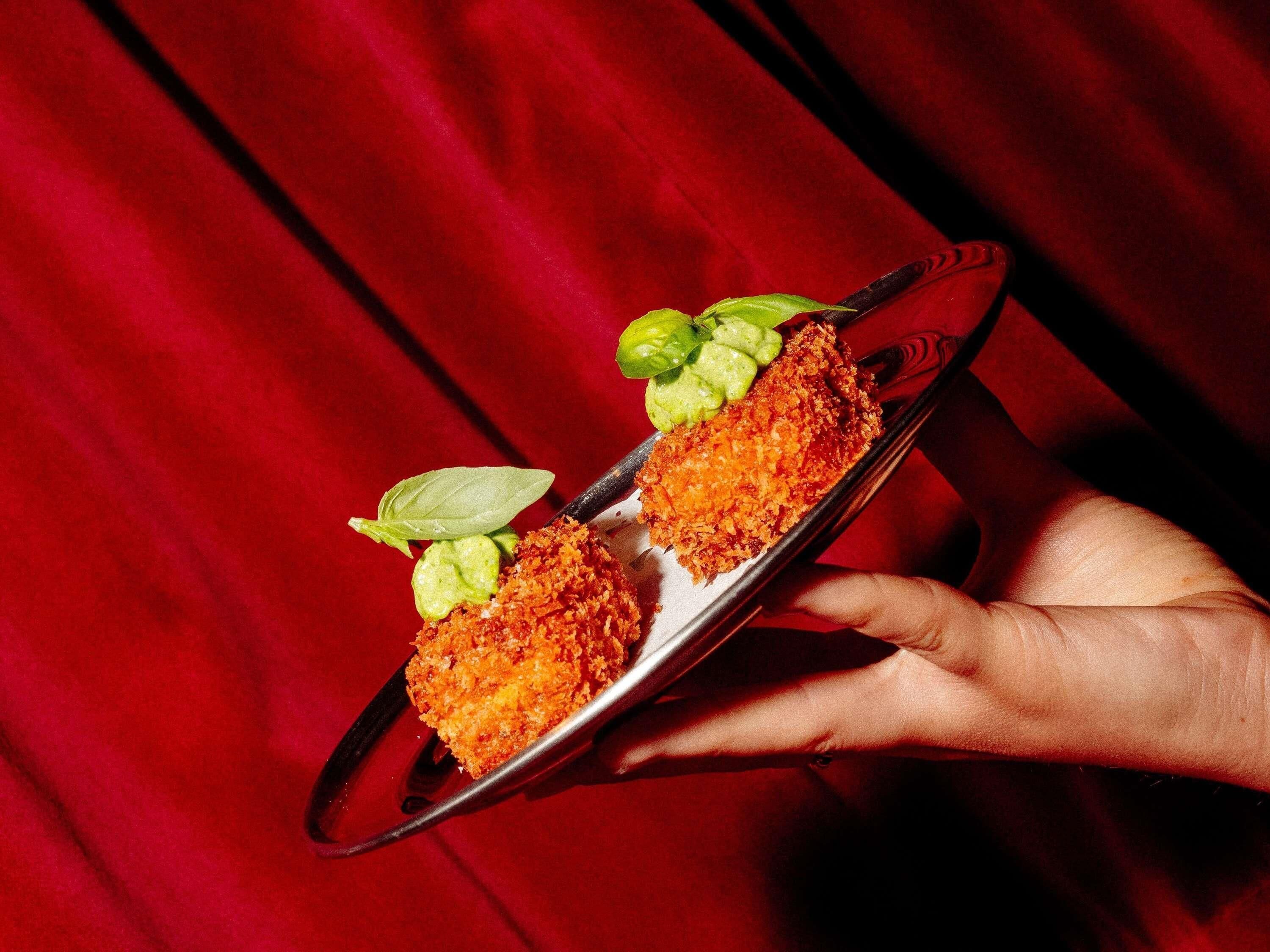 A hand holding a plate with a food item, possibly a breaded cutlet, garnished with a green leaf and a green sauce.