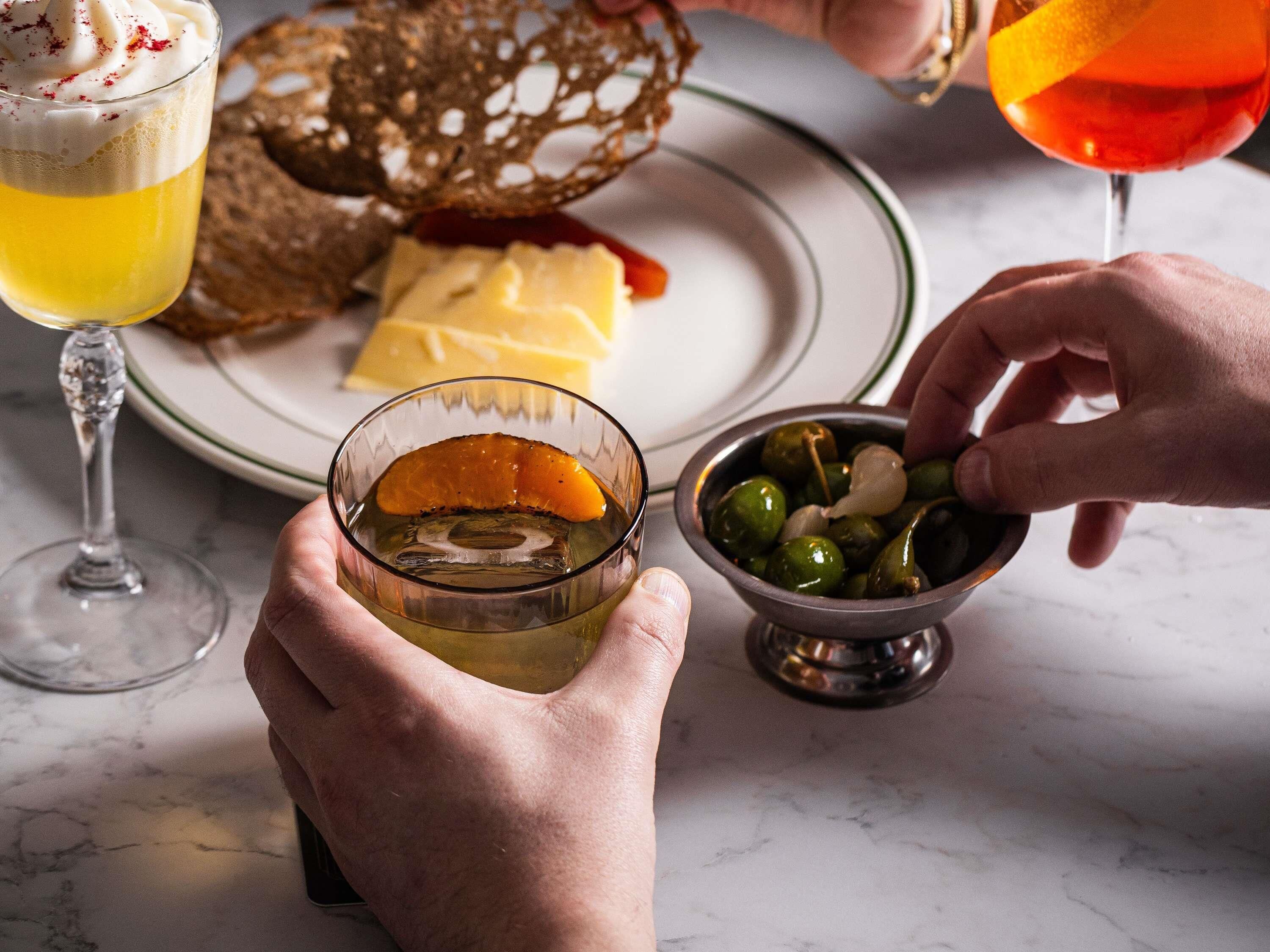 A person holding a glass with a yellow beverage, a bowl of olives, and a plate with bread and cheese.