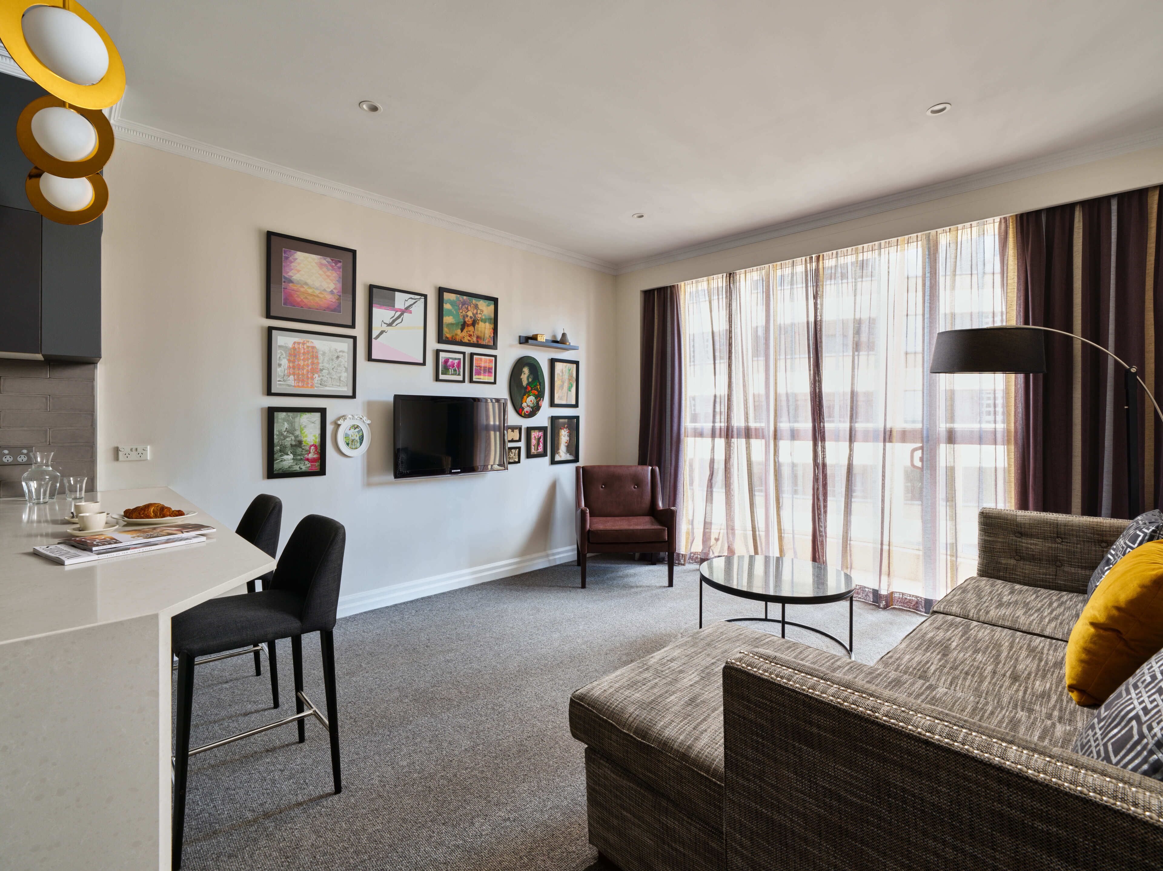 A modern living room with a sofa, a dining table, and a TV. There are framed pictures on the wall and a window with curtains.
