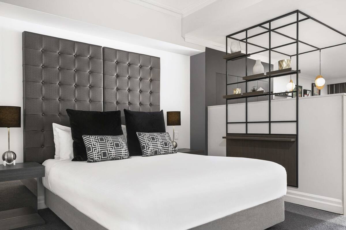 A modern bedroom with a tufted headboard, a bed with white bedding, black and white patterned pillows, a nightstand with a lamp, and a built-in shelving unit with decorative items.