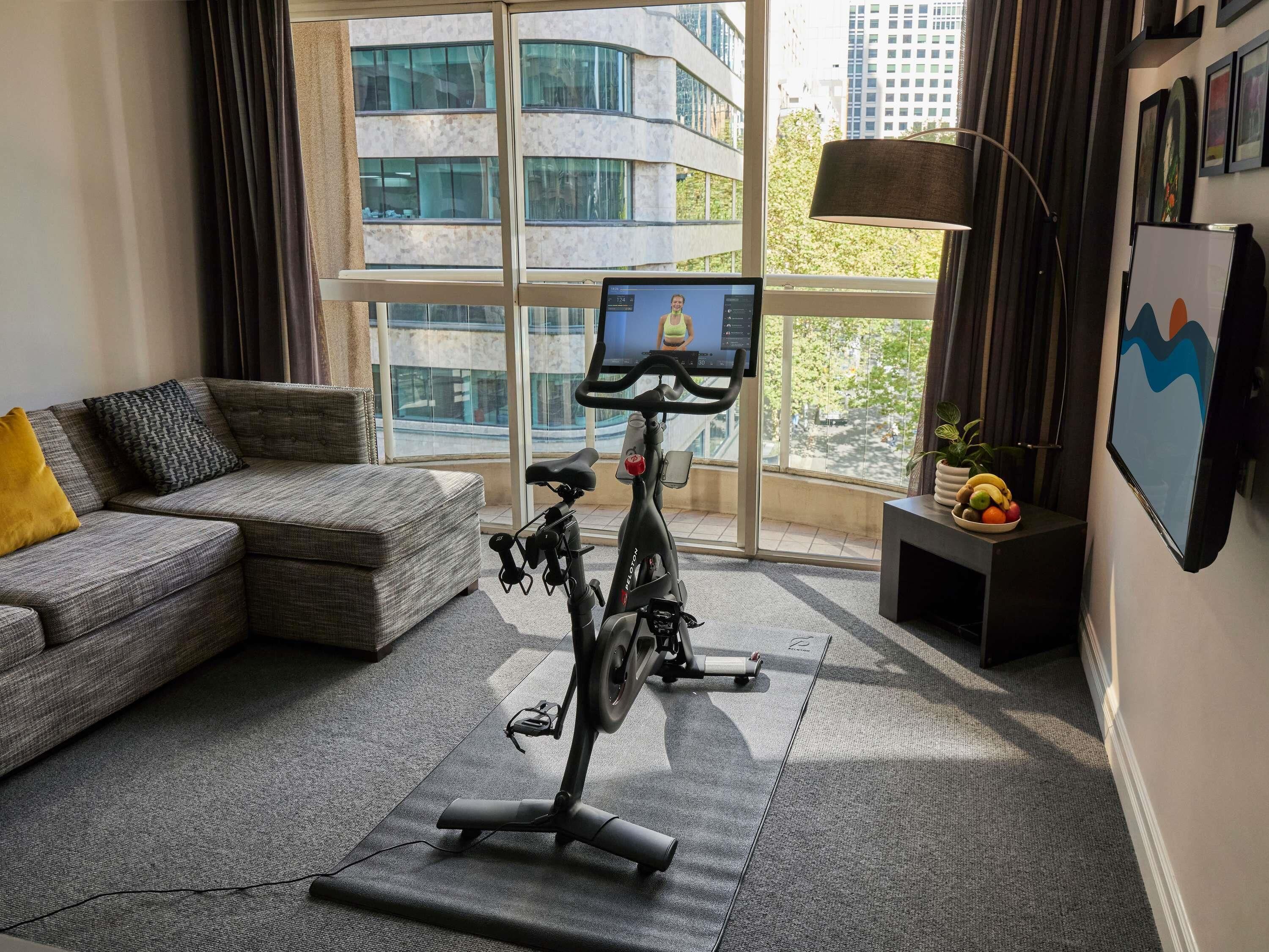 Ein modernes Wohnzimmer mit einem grauen Sofa, einem Steh-Trainingsrad, einem kleinen Tisch mit Obst und einem großen Fenster mit Blick auf die Stadt.