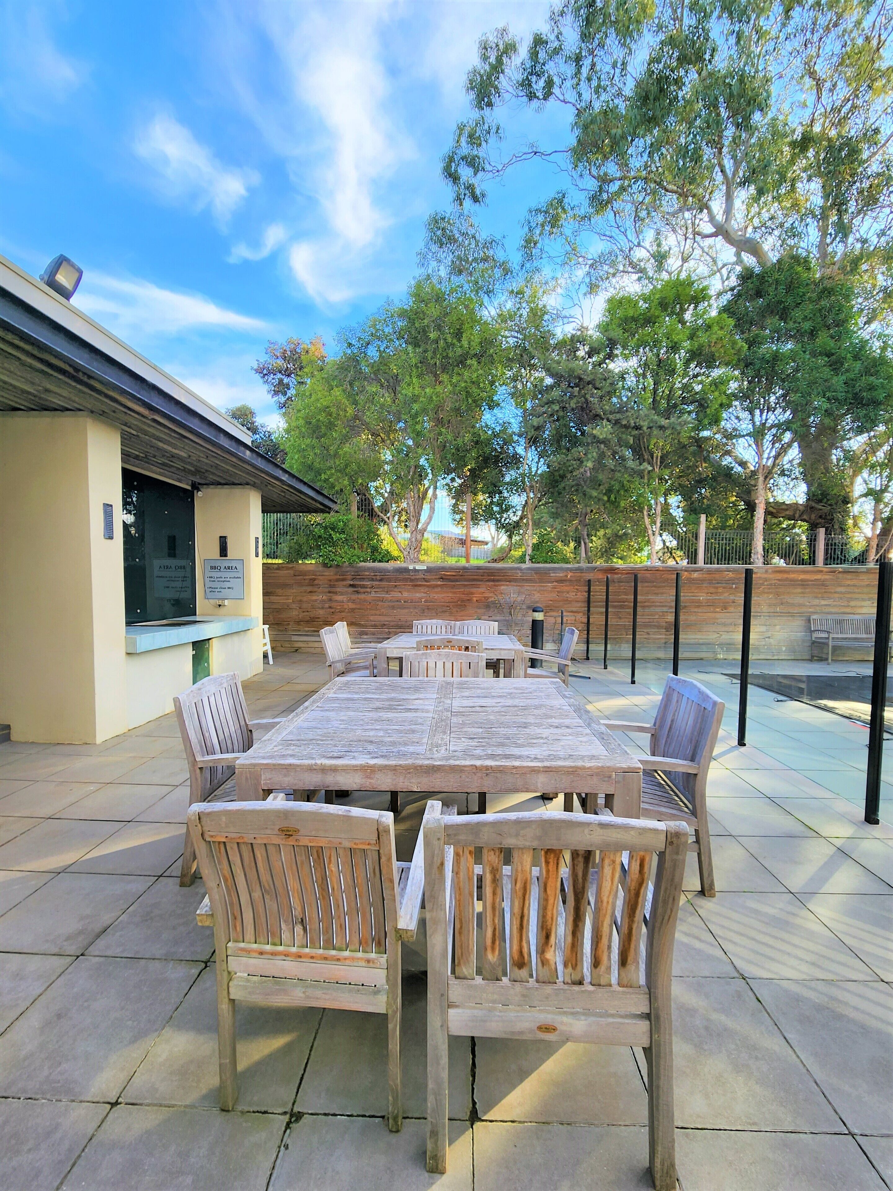 Un patio al aire libre con mesa y sillas de madera, un letrero que indica área de barbacoa y vistas a árboles y una cerca.