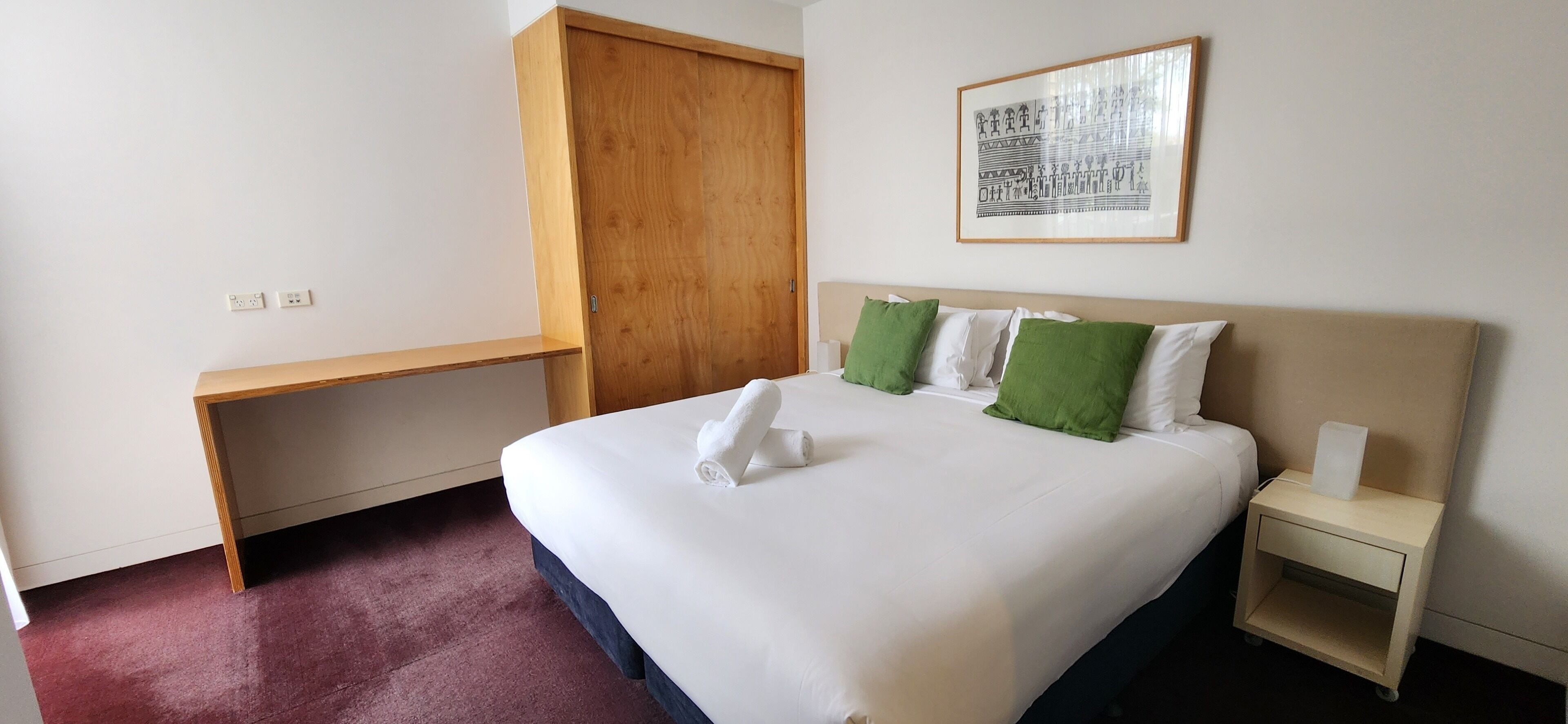 A hotel room with a bed, a wooden desk, a nightstand, and a wall-mounted artwork.