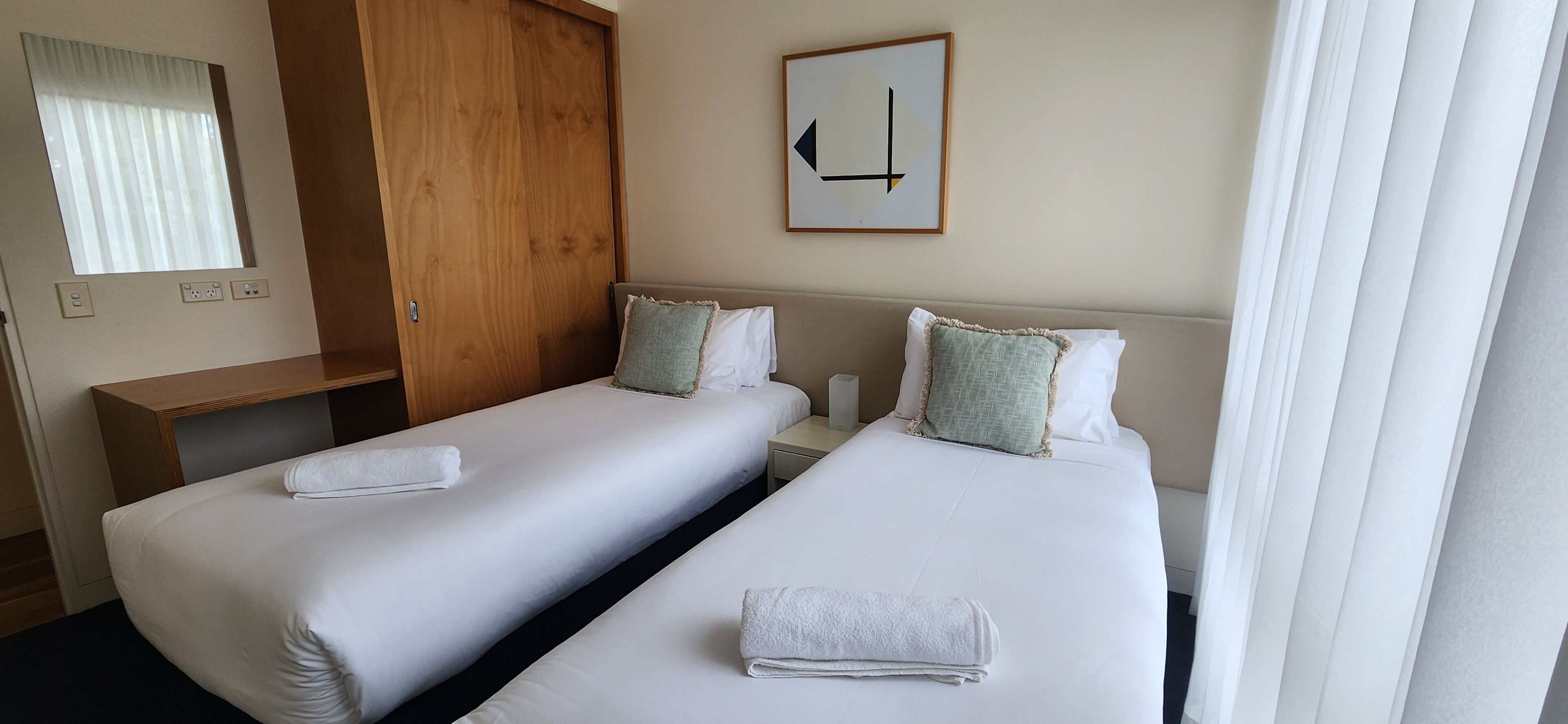 A hotel room with two beds, a wooden wardrobe, a mirror, and a framed picture on the wall.
