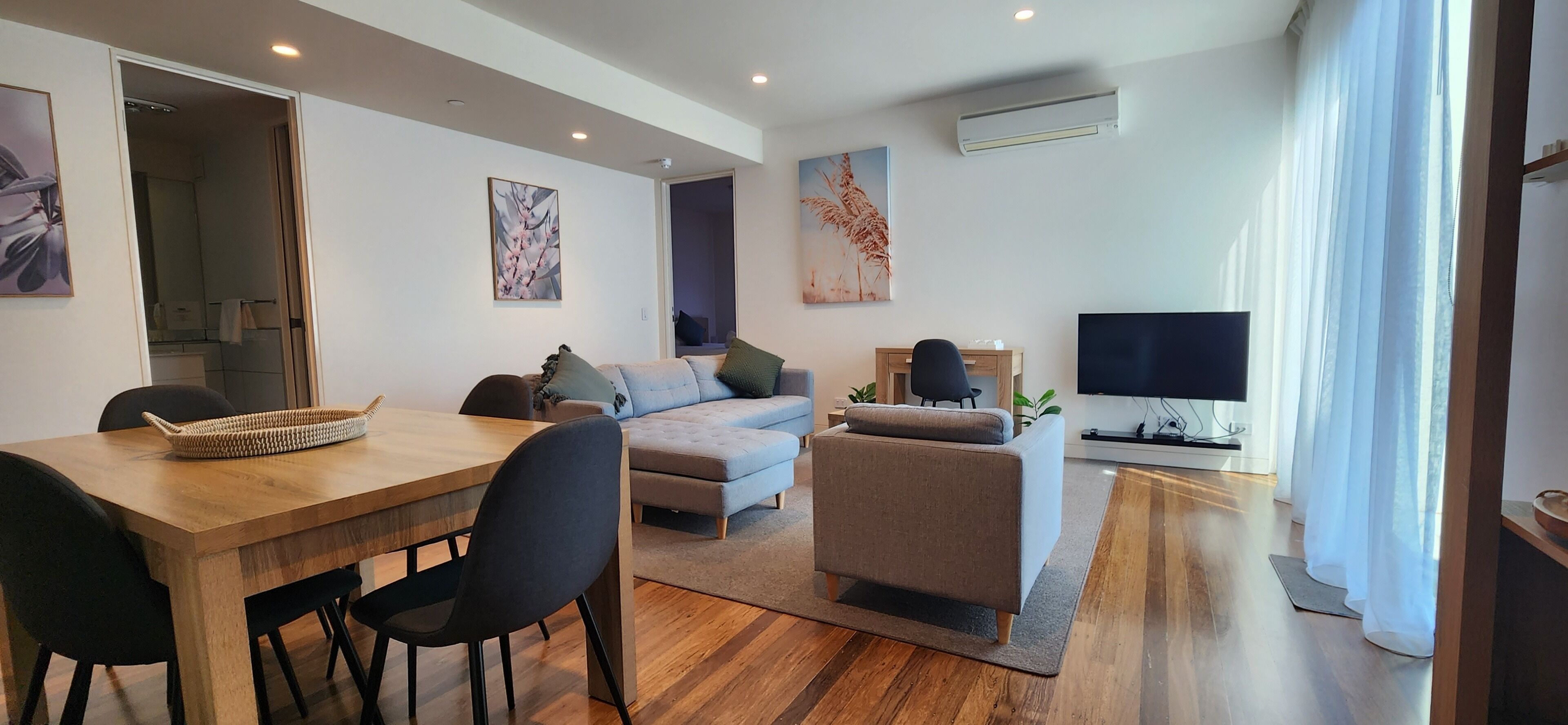 A modern living room with a wooden dining table, a grey sofa, a grey armchair, and a flat-screen TV.