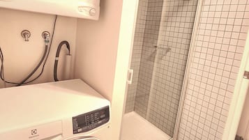 Un cuarto de lavado con lavadora y secadora de carga frontal, un área de ducha con azulejos y la instalación de fontanería a la vista.
