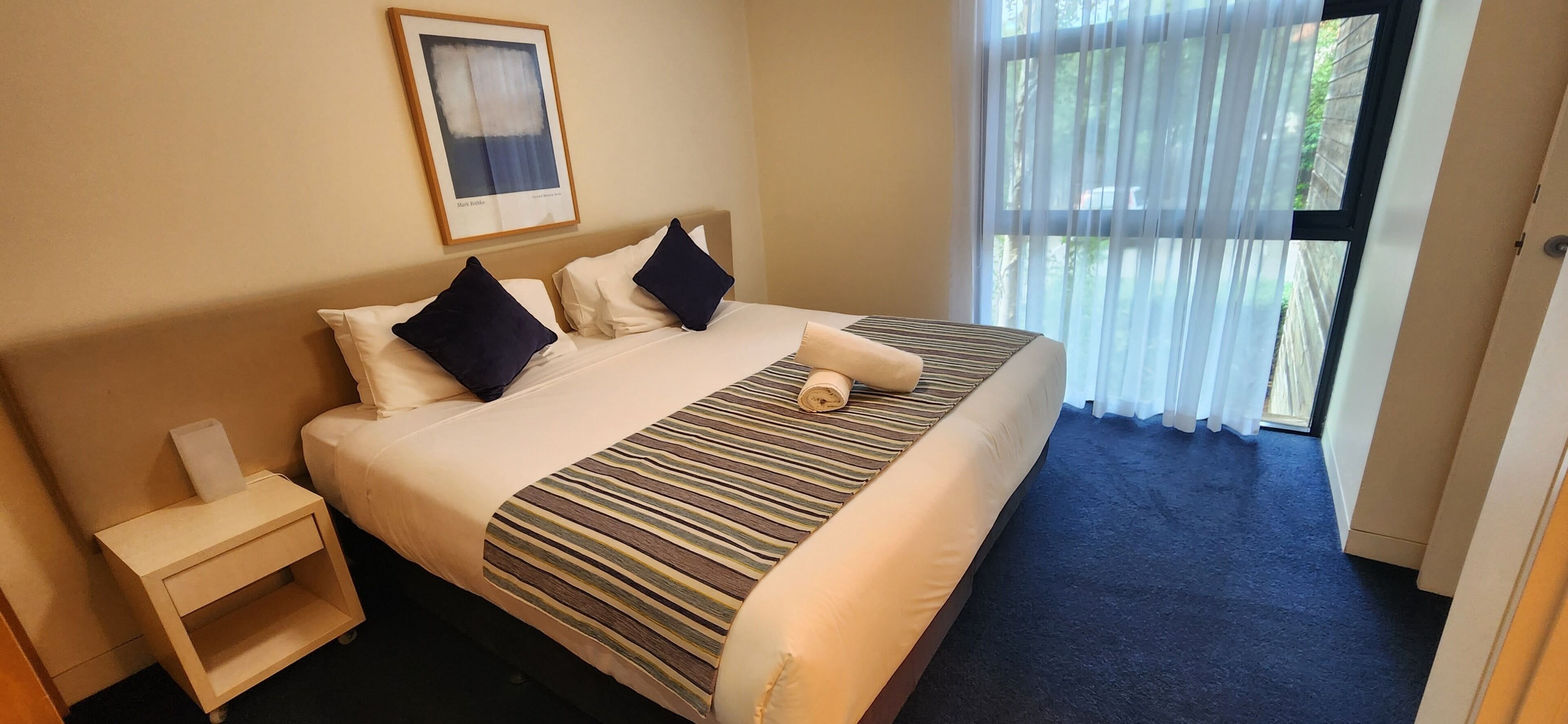 Ein Hotelzimmer mit Bett, Nachttisch, einem gerahmten Bild an der Wand und Blick ins Freie durch ein Fenster.