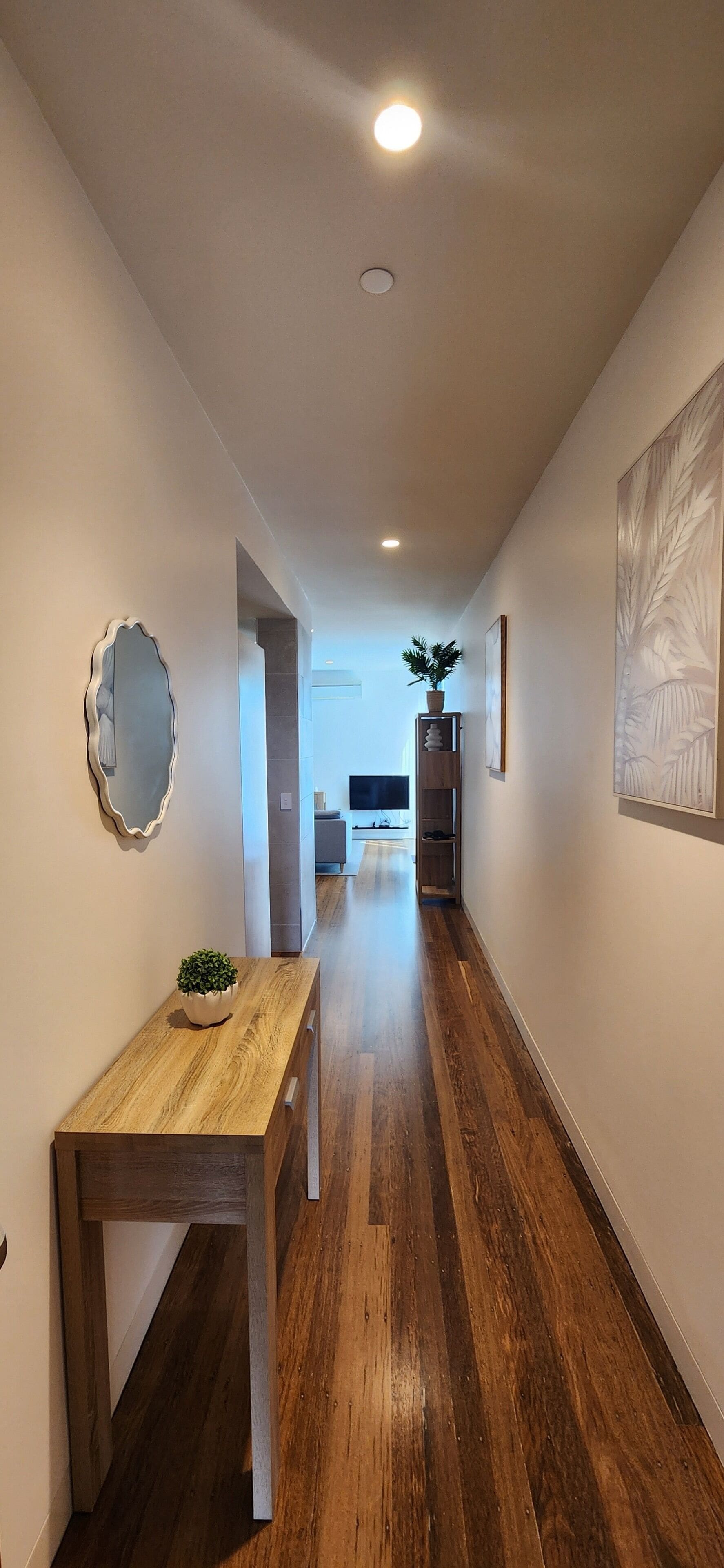 A hallway with wooden flooring, a small table with a plant, a mirror, and a framed picture on the walls.