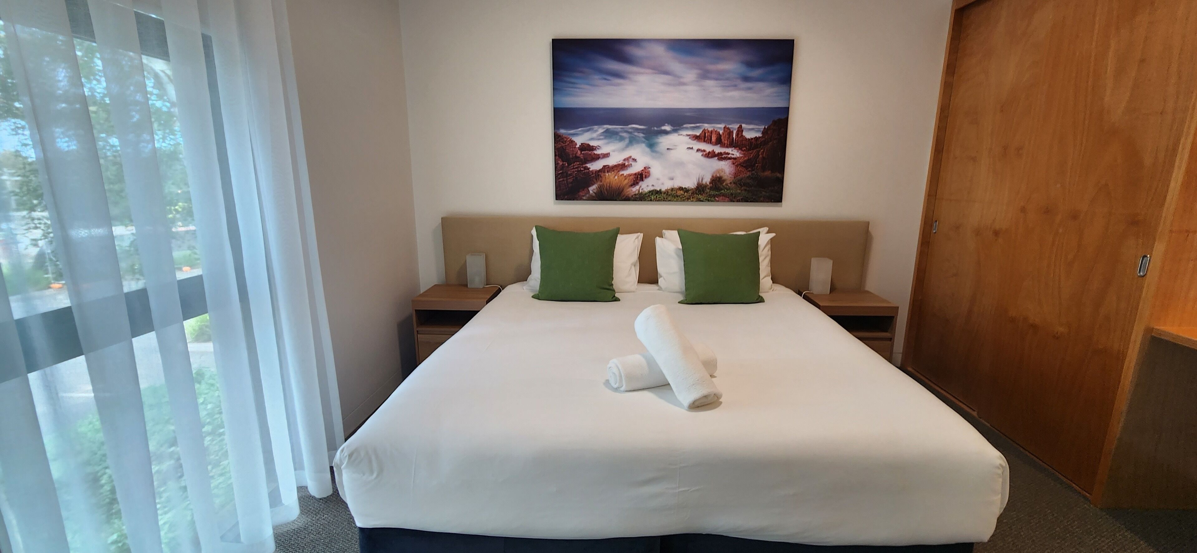 Habitació d'hotel amb un llit gran, dues mesetes de nit, un armari de fusta i un quadre d'un paisatge costaner.
