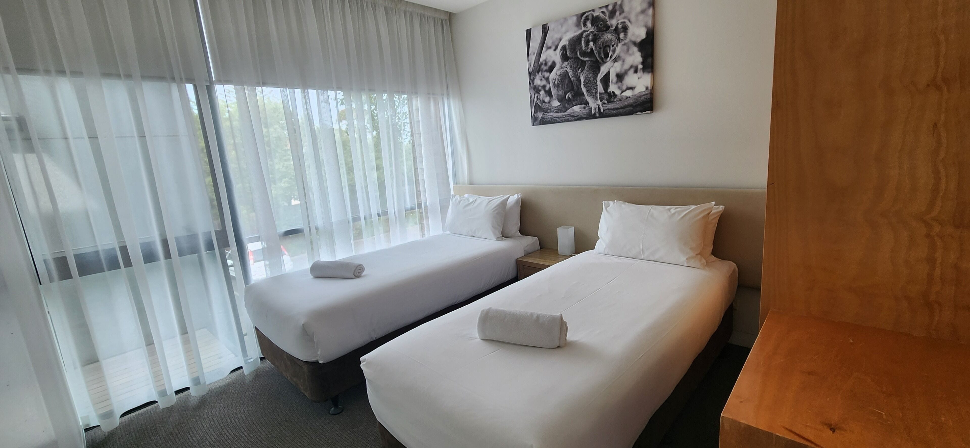 Ein Hotelzimmer mit zwei Einzelbetten, einem großen Fenster mit transparenten Vorhängen, einem hölzernen Kopfteil und einem gerahmten Bild an der Wand.
