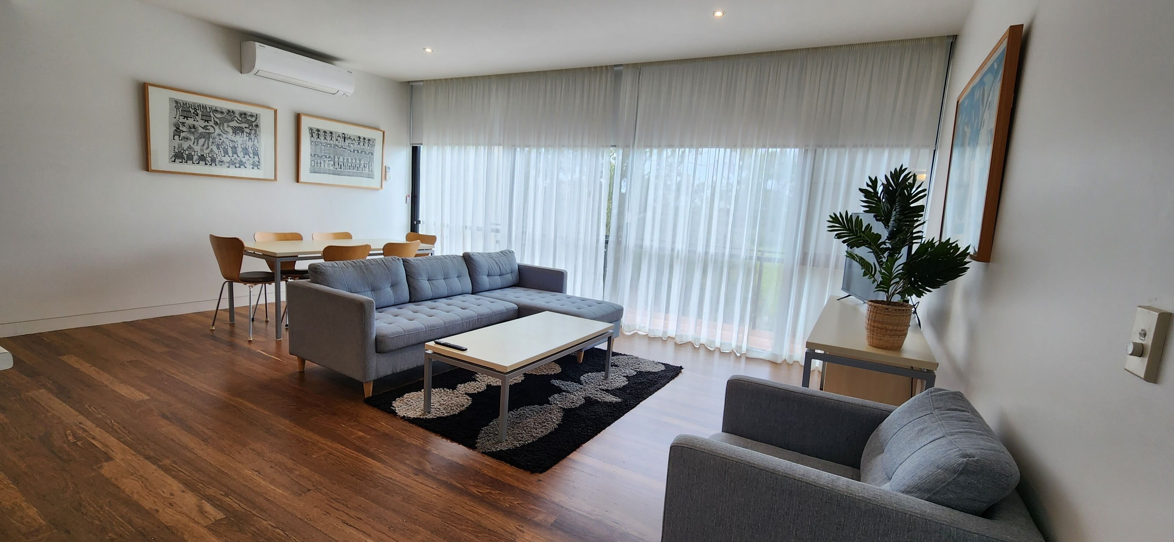 A modern living room with a grey sectional sofa, a small table, and a coffee table. There are two chairs, a potted plant, and artwork on the walls.
