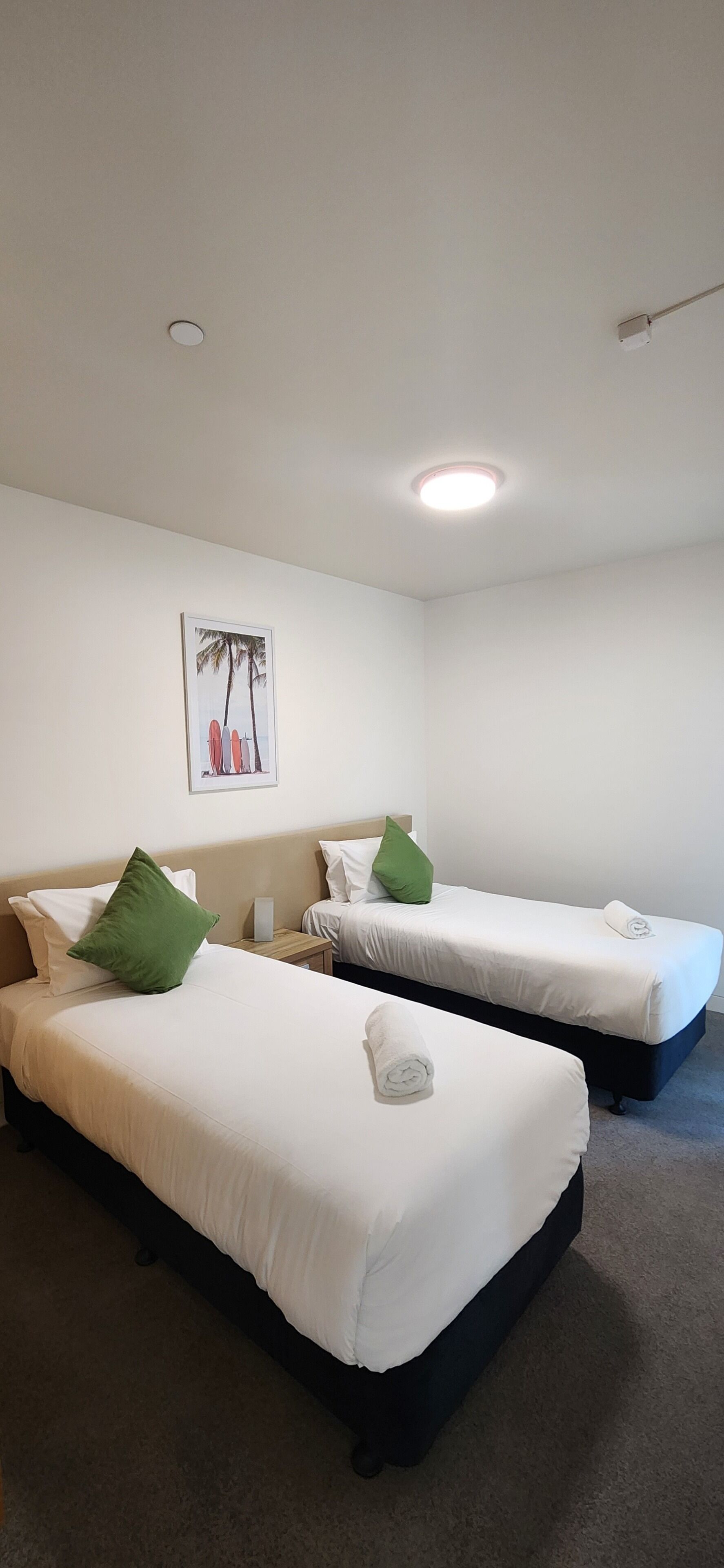 A hotel room with two single beds, each with white bedding and green pillows. There is a ceiling light and a framed picture of surfboards on the wall.
