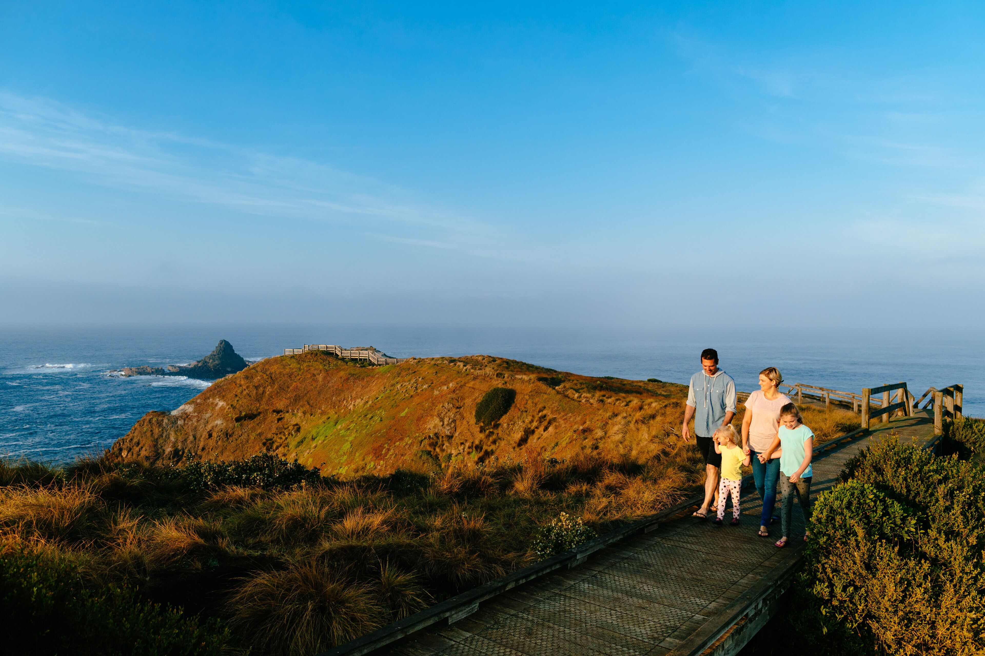 A family on a wooden boardwalk overlooking a coastal landscape with a rocky hill and ocean view.