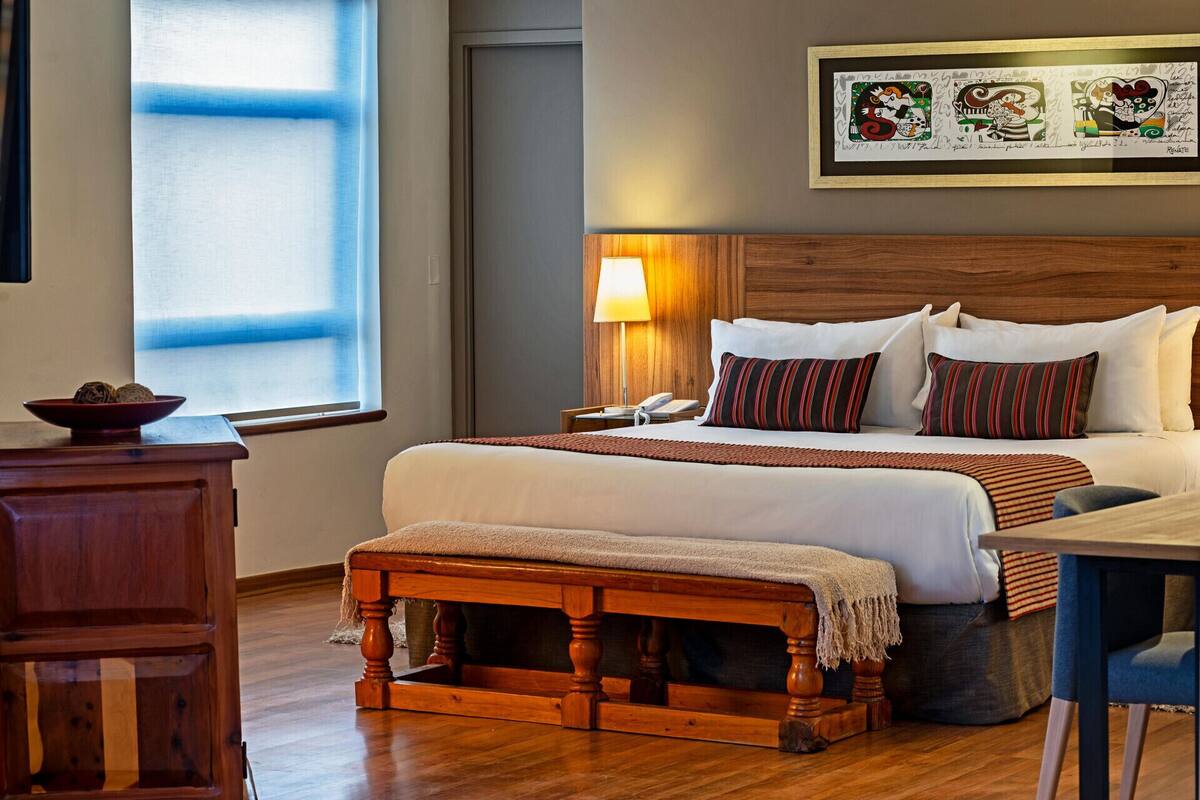 A hotel room with a wooden headboard, a bed with white bedding, striped pillows, a bedside lamp, a wooden bench, a desk, and a framed artwork on the wall.