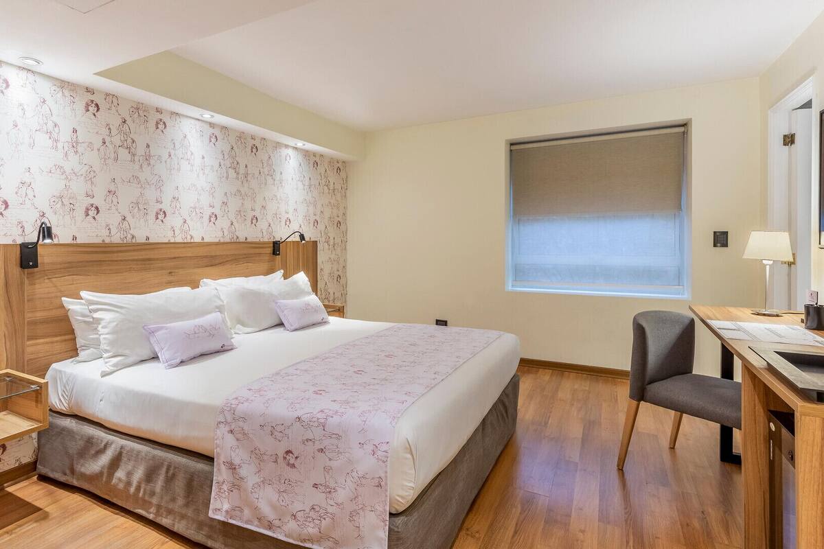 A hotel room with a bed, desk, chair, and a window with blinds.