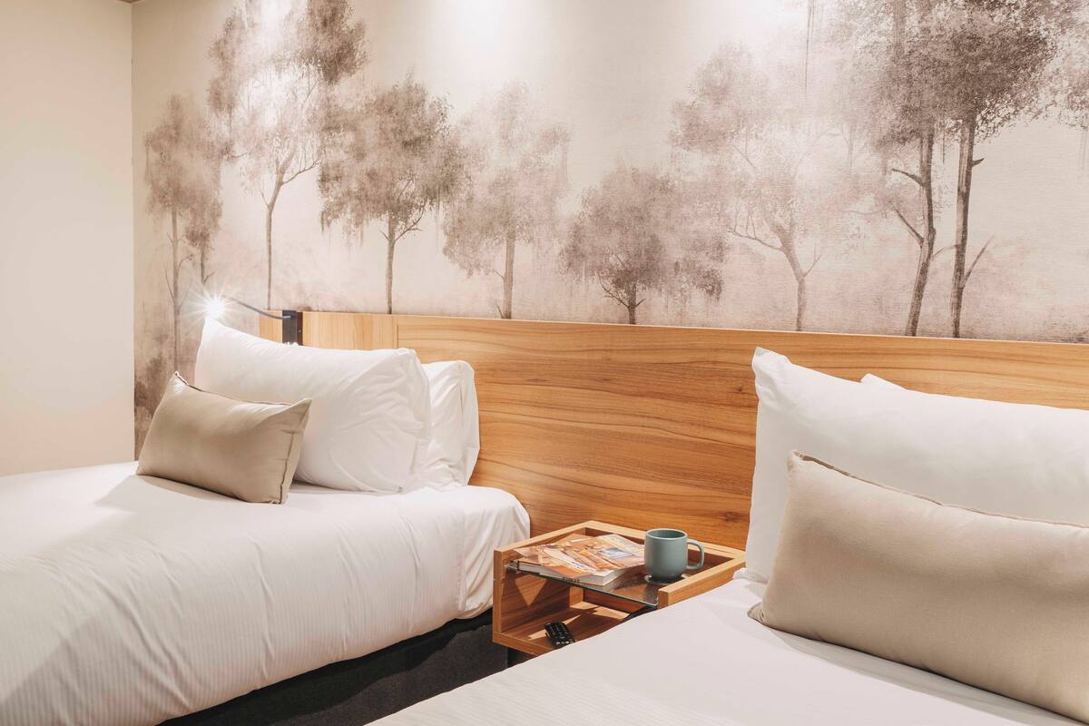 A hotel room with two beds, a wooden headboard, a nightstand with a tray holding a book and a mug, and a wall mural of trees.