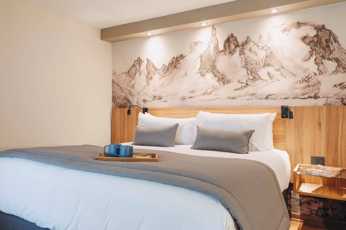 A neatly made bed with a tray of items, a wooden headboard, and a wall with a mountain painting.