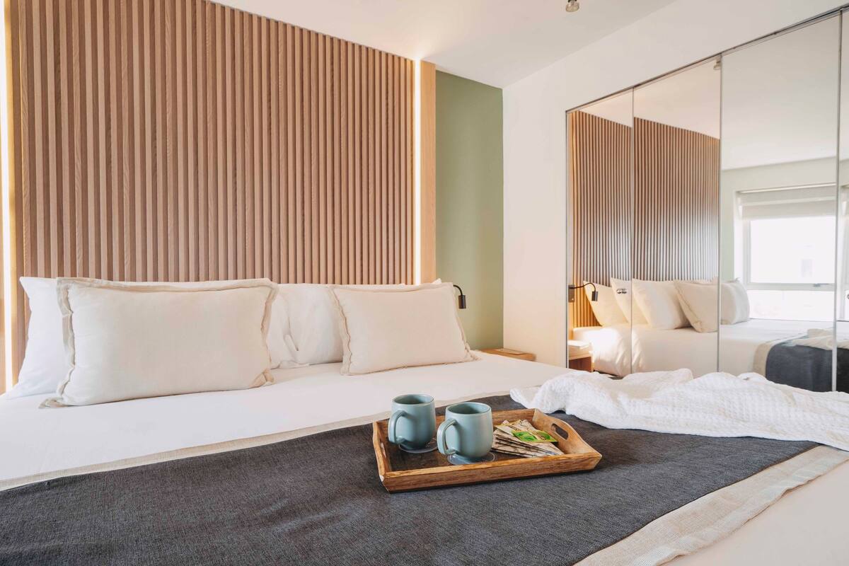 A modern hotel room with a large bed, a wooden headboard, and a tray with two mugs and a book.