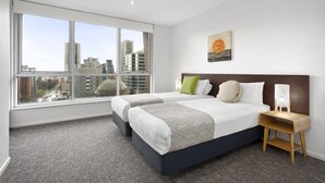 Habitación de hotel con dos camas, un ventanal con vista a la ciudad, una mesita de noche con lámpara y una obra de arte colgada en la pared.