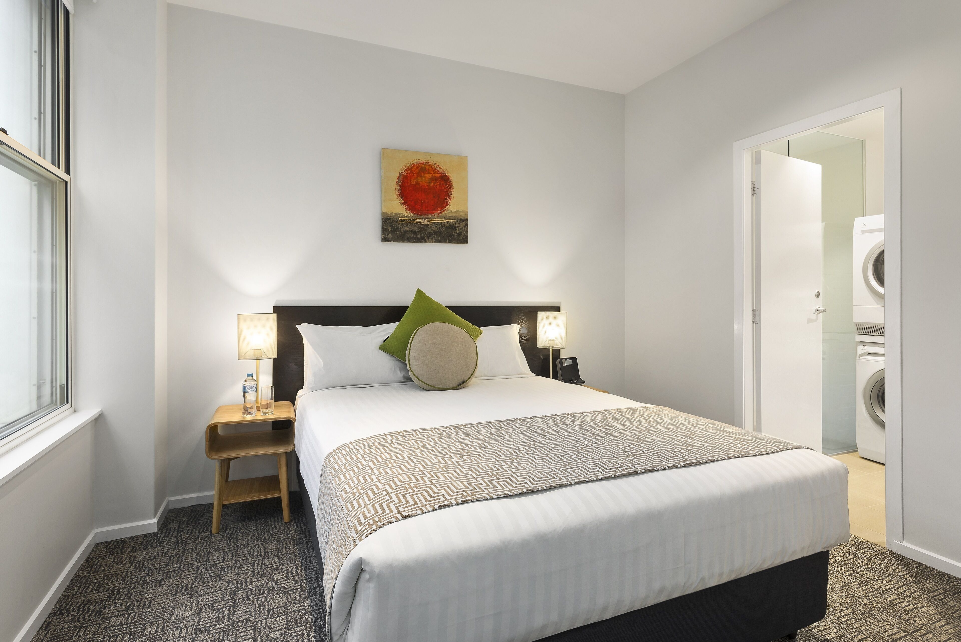 A hotel room with a neatly made bed, a nightstand with a lamp, a painting on the wall, and a washing machine in the corner.