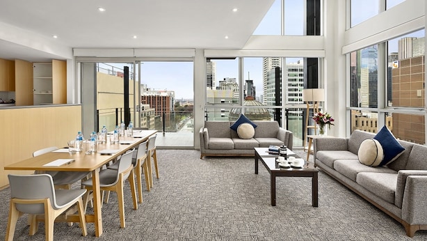 Habitación de hotel moderna con comedor, un conjunto de sofás y ventanas amplias con vistas a la ciudad.