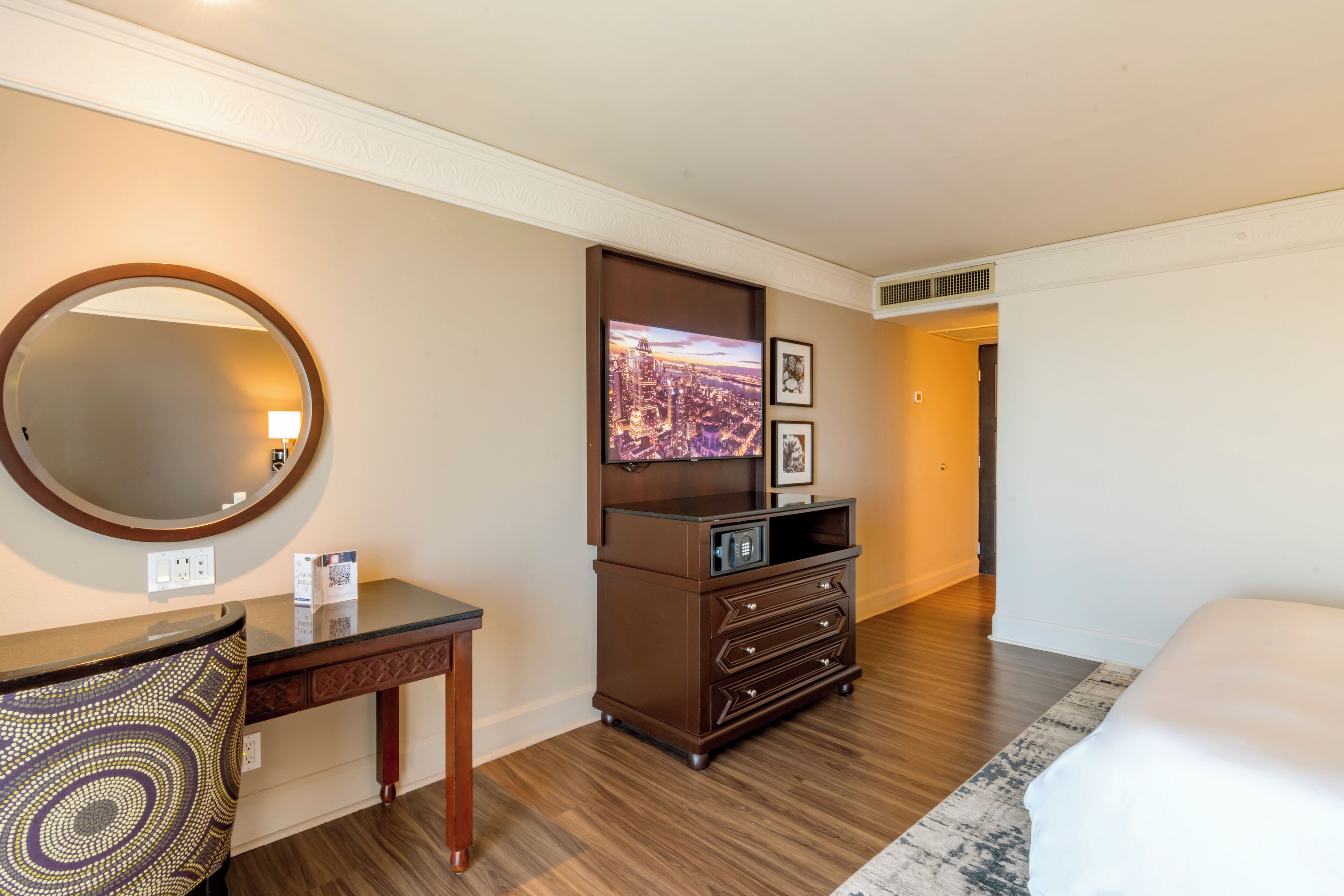 Una habitación de hotel con piso de madera, un espejo grande, un televisor de pantalla plana, un aparador y una cama.