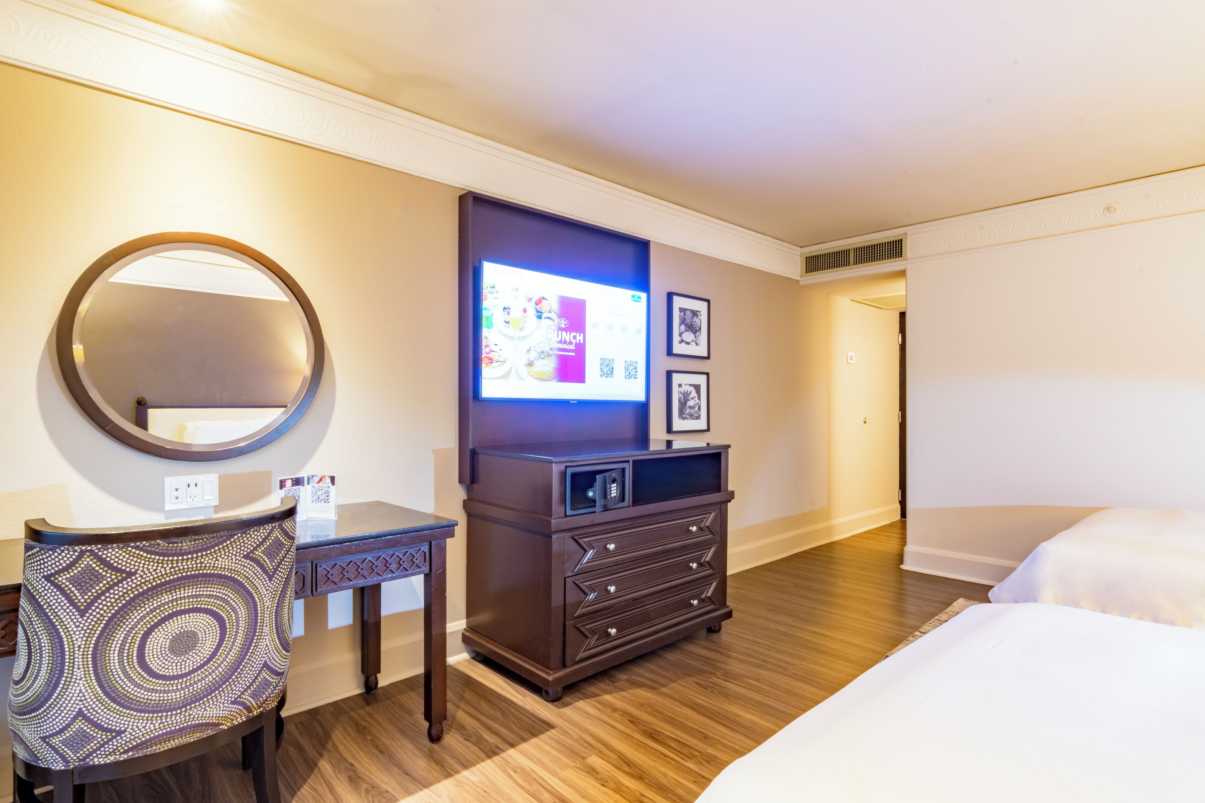 A hotel room with a bed, a desk with a chair, a dresser, a TV, and framed pictures on the wall.
