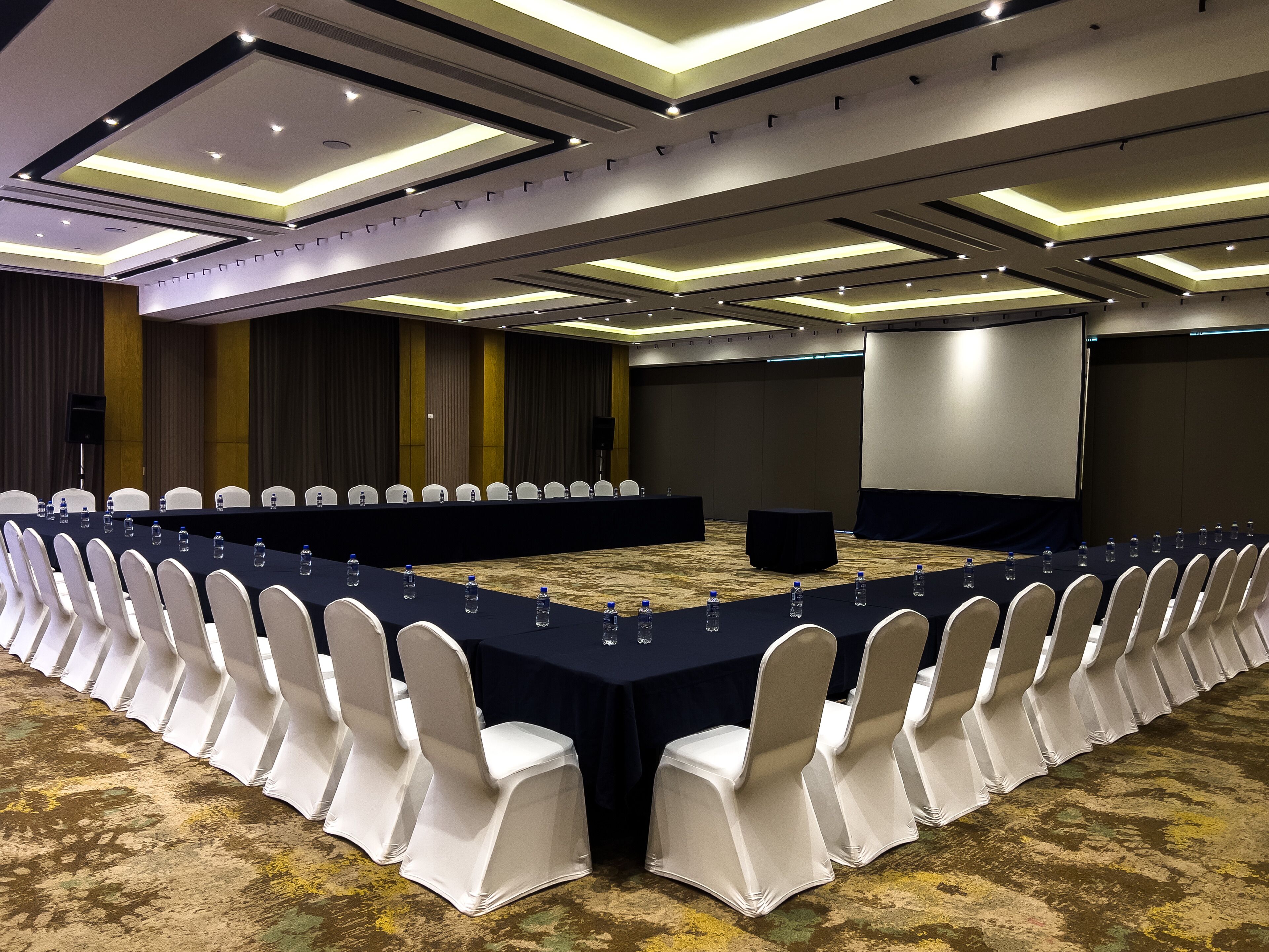 Un salón de conferencias con una larga mesa, sillas blancas y una pantalla para proyector.