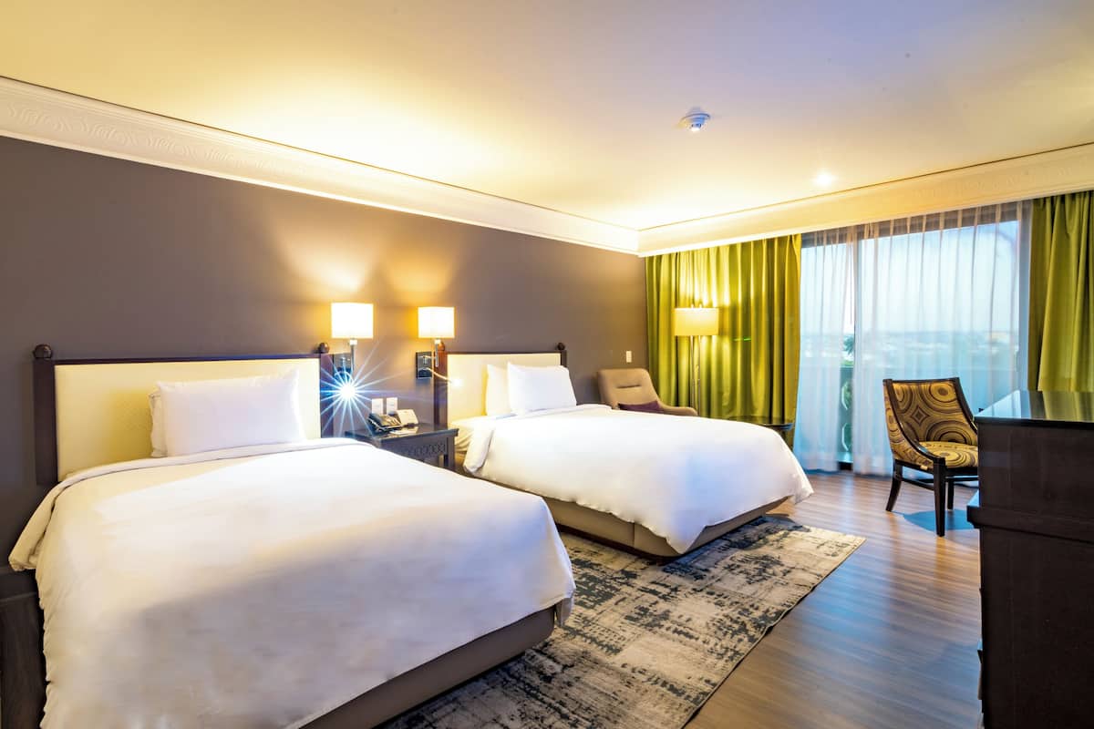 A hotel room with two beds, a desk, a chair, and a view of the city.