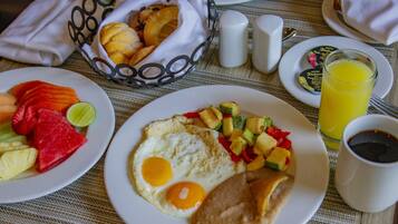 Desayuno con platos de huevos, fruta y tostadas, una cesta de bollería y un vaso de zumo de naranja.
