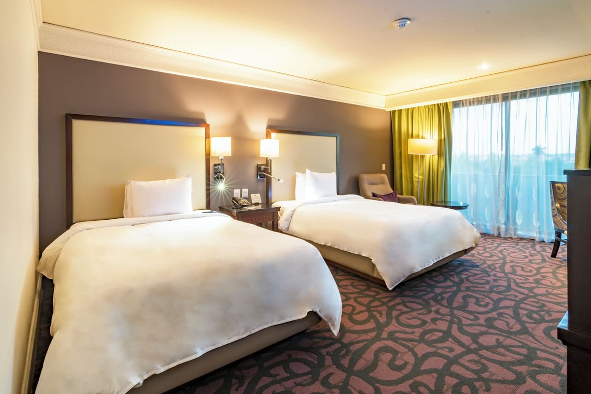 A hotel room with two beds, a large mirror, bedside lamps, a desk, and a view of the city through large windows.