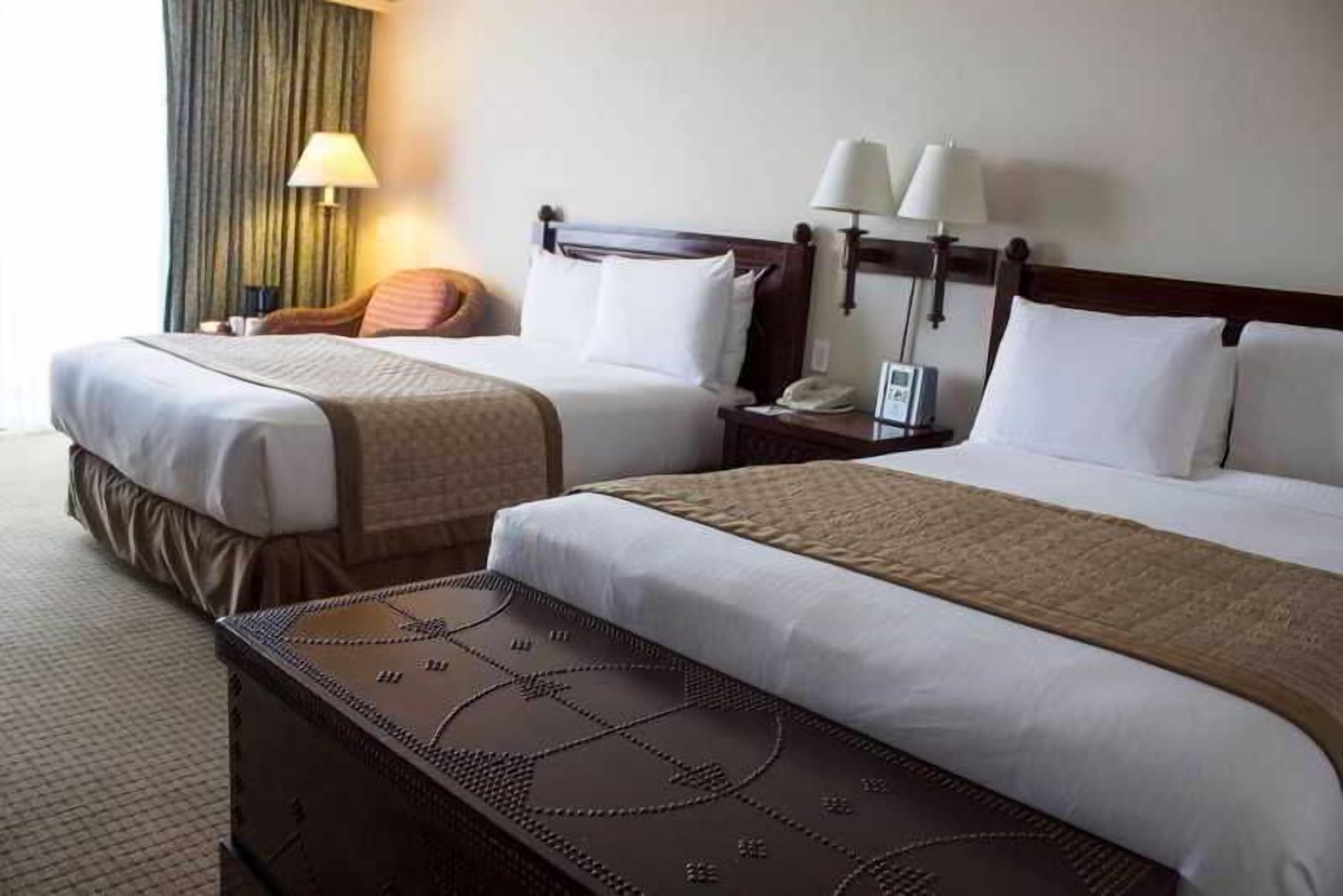 A hotel room with two beds, a nightstand with a lamp, and a chair.