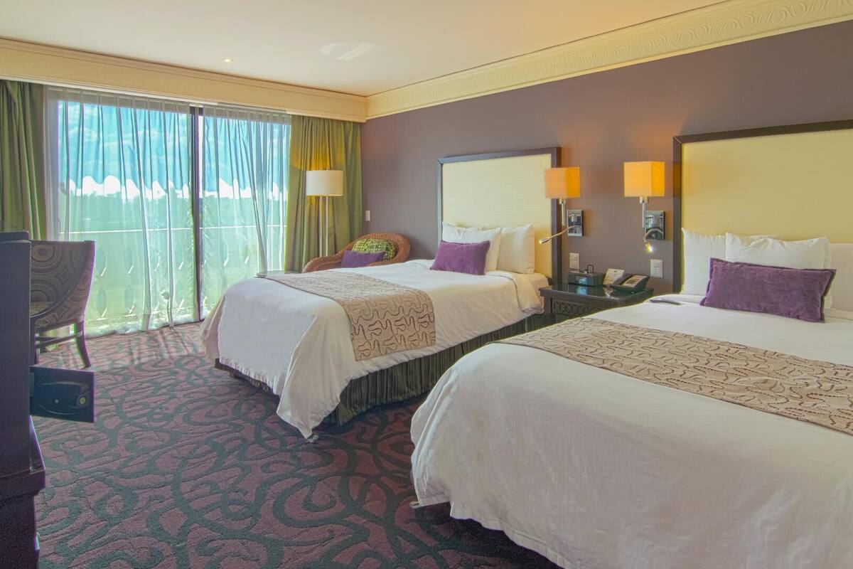 A hotel room with two beds, a chair, a desk, and a large window with curtains.