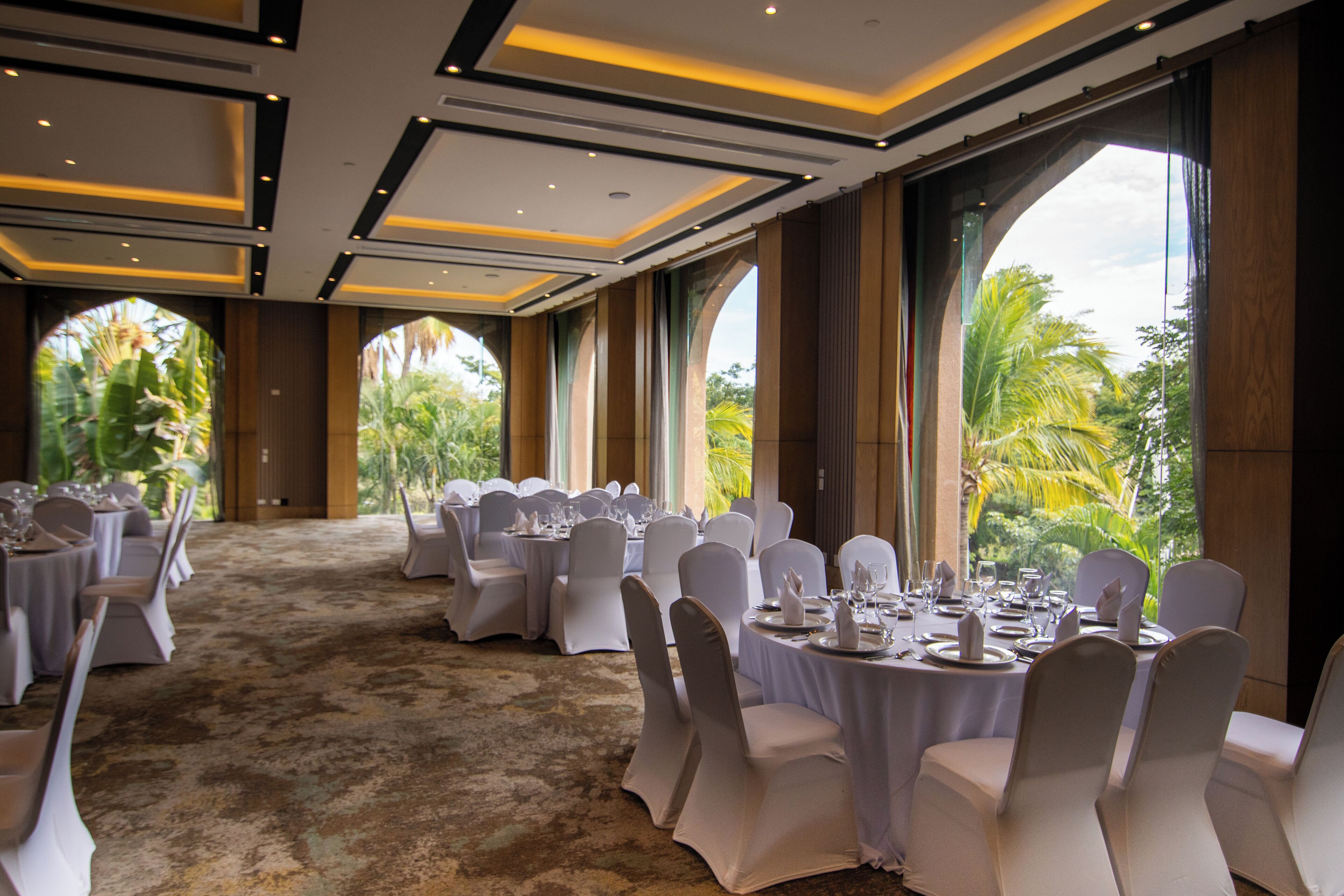 Un salón de banquetes con mesas redondas dispuestas para comer, amplias ventanas con vistas a la vegetación y suelo cubierto de alfombra.