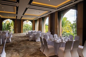 Un salón de banquetes con mesas redondas dispuestas para comer, amplias ventanas con vistas a la vegetación y suelo cubierto de alfombra.